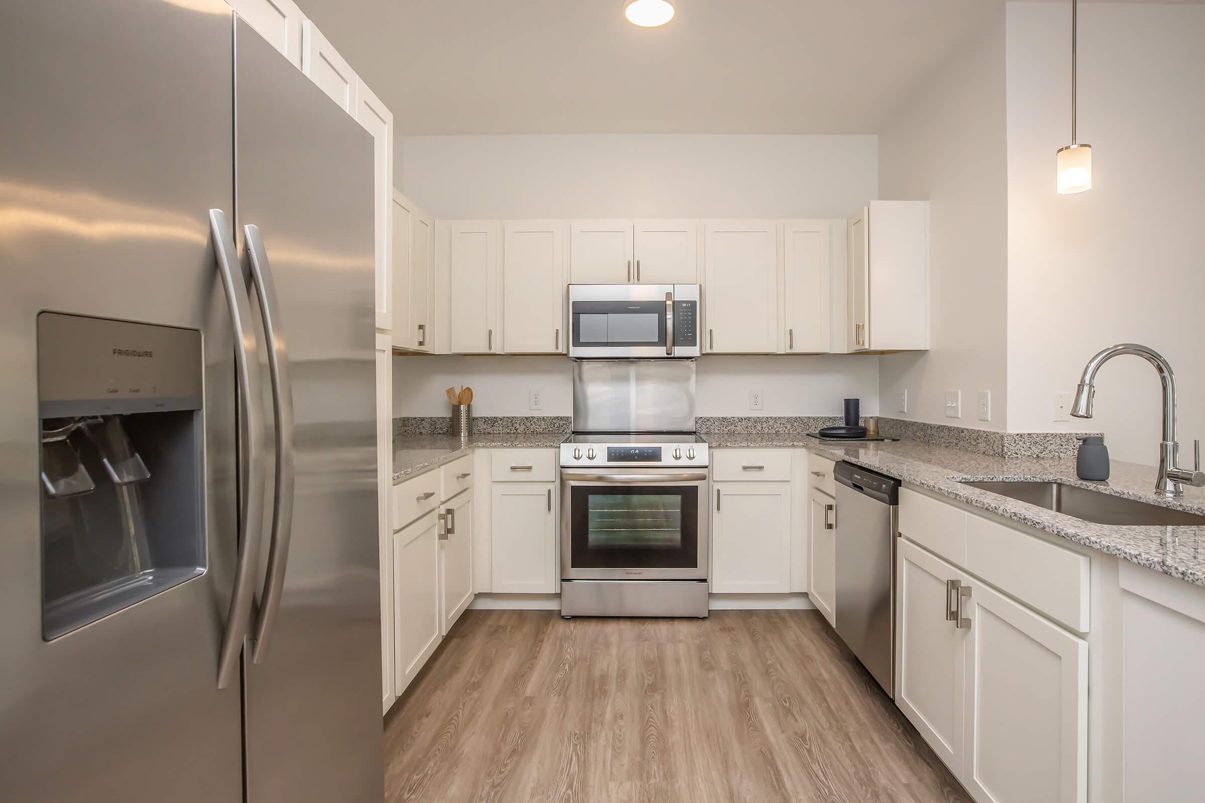 Modern kitchen with stainless steel appliances, including a refrigerator, oven, and microwave. White cabinets and a granite countertop create a bright and clean look. The kitchen features a sink and a pendant light above, with light wood flooring enhancing the contemporary design.