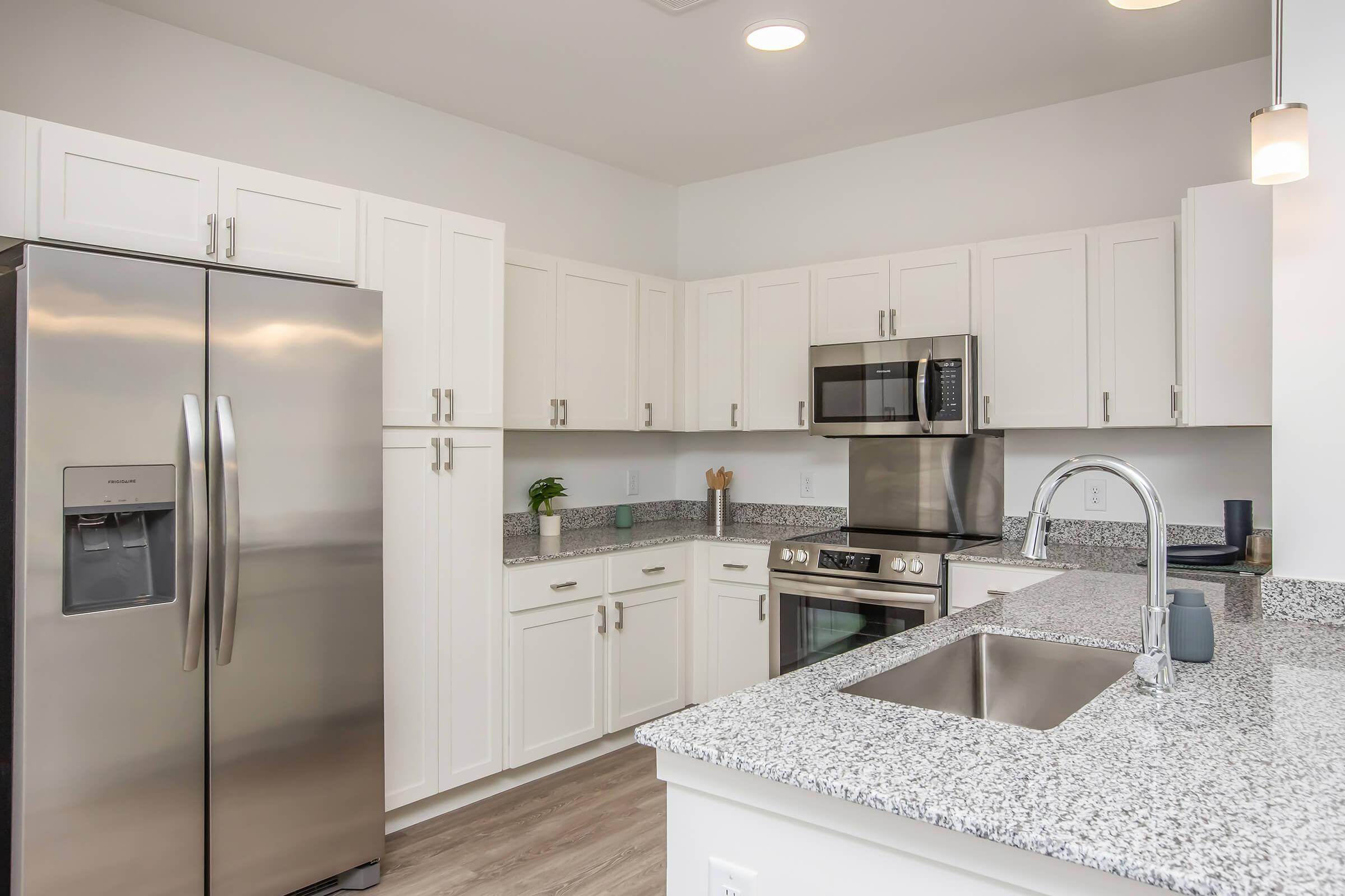 A modern kitchen featuring stainless steel appliances, including a refrigerator and microwave, granite countertops, and white cabinetry. The space is bright with recessed lighting and a minimalist design, showcasing a clean and open layout perfect for cooking and entertaining.