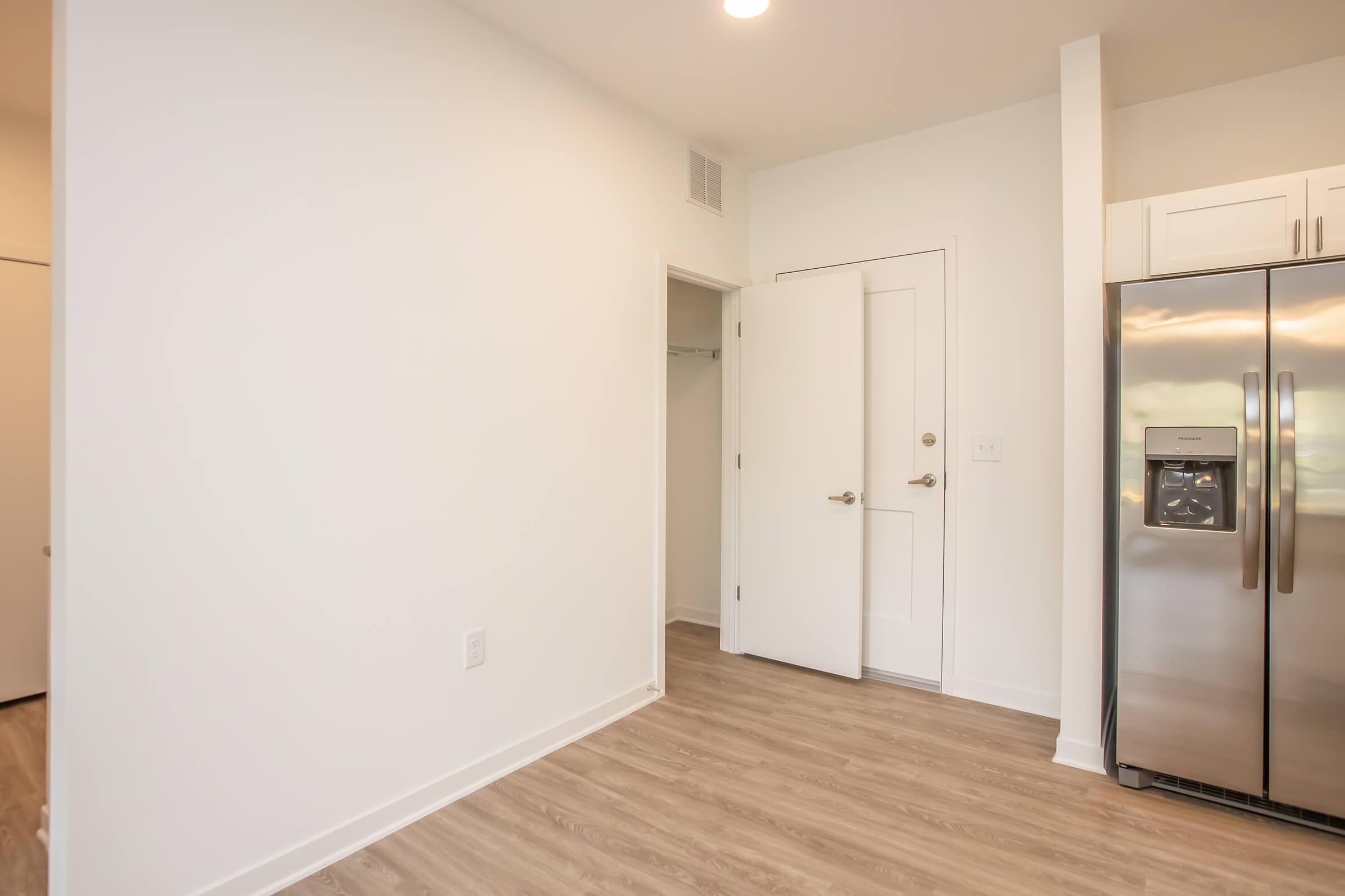 Interior view of a modern kitchen area, featuring a stainless steel refrigerator on the right side, light wood-style flooring, and white walls. A doorway leads to a closet and another room. The space is well-lit with natural light and has a clean, minimalistic design.