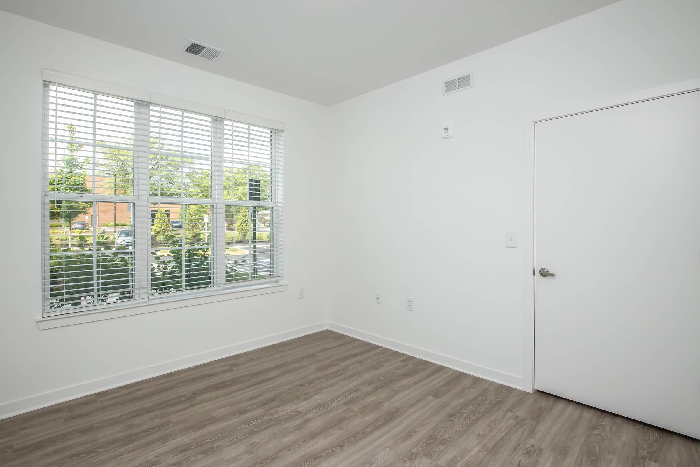 Empty room with light-colored walls, featuring a large window with white blinds allowing natural light to enter. The floor is finished with light wood-like laminate. A door is visible on one side, and there are no furnishings or decorations in the space, giving it a clean and modern appearance.