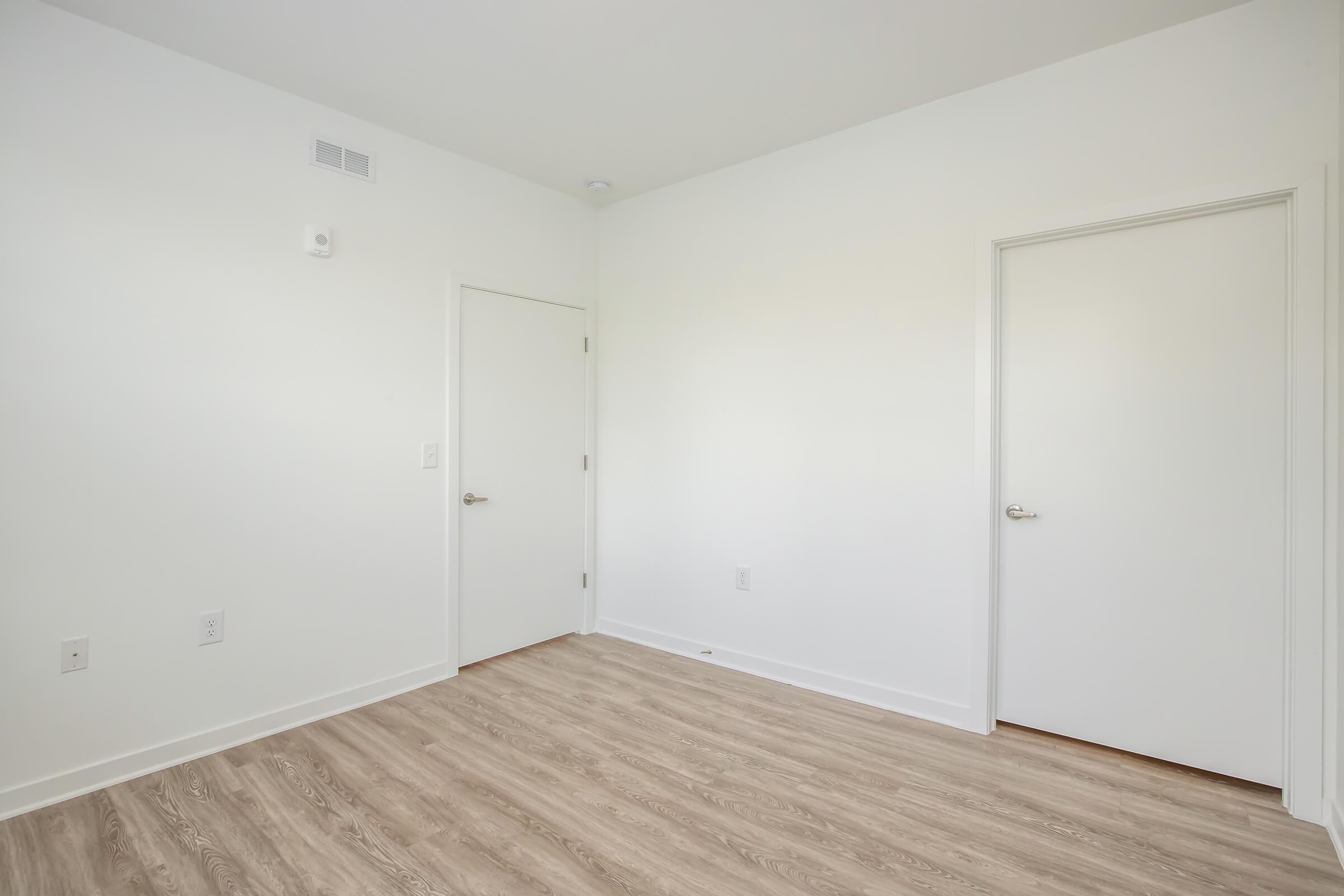 Empty room with light-colored walls and two closed doors. The floor is covered in light wood laminate. There are no furnishings or decorations in the space, giving it a clean and minimalistic appearance. Natural light from a window may be present but is not visible in the image.