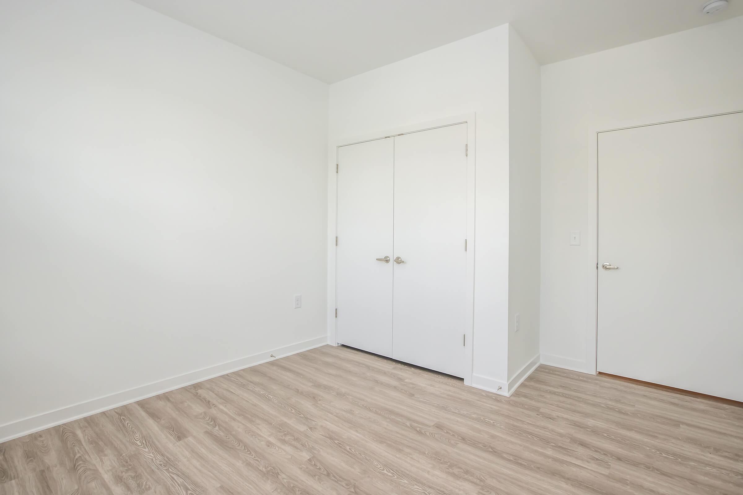 A bright, empty room with light-colored walls and a light wood floor. Two closed white closet doors are on the left side, and a single door is on the right. The space is uncluttered, emphasizing its spaciousness and simplicity, ideal for a fresh start or new furnishings.