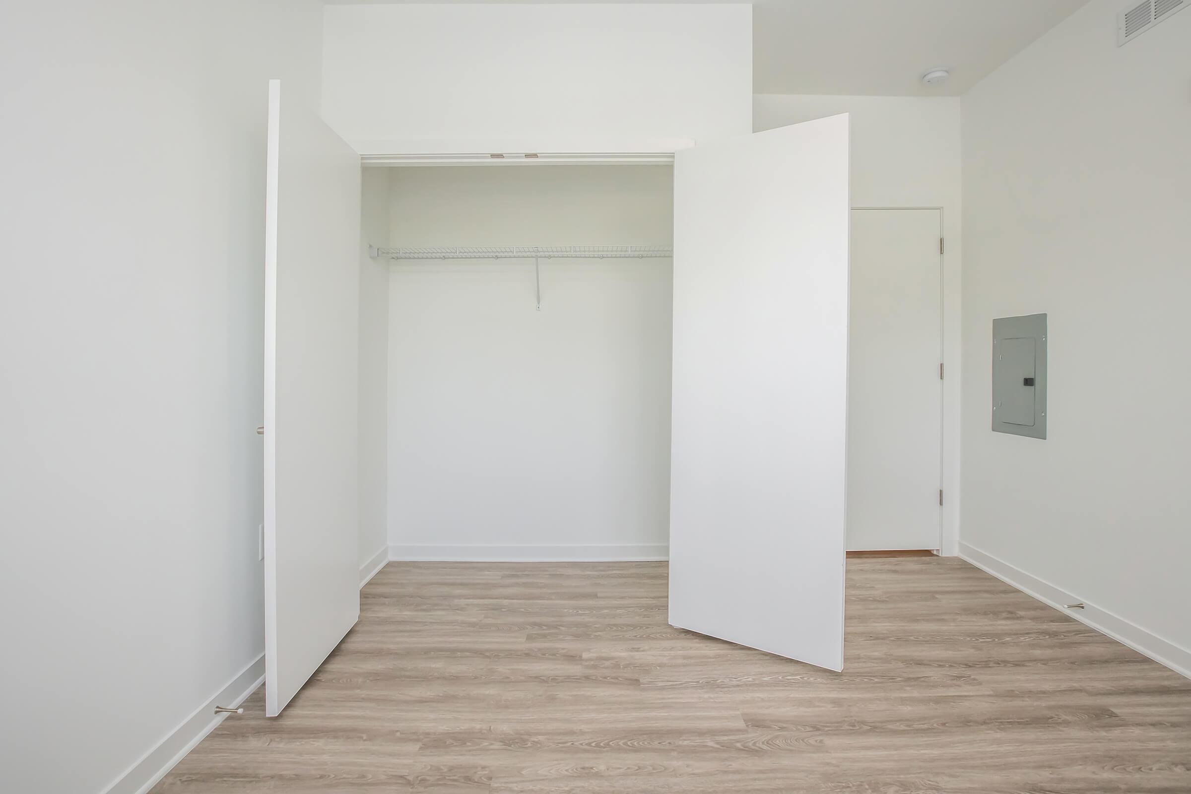 Empty room featuring a spacious closet with open double doors, a single hanging rod, and a light-colored floor. The walls are plain and painted in a soft white, creating a bright and minimalist atmosphere. A door is visible to the right, with an electrical panel mounted on the wall.