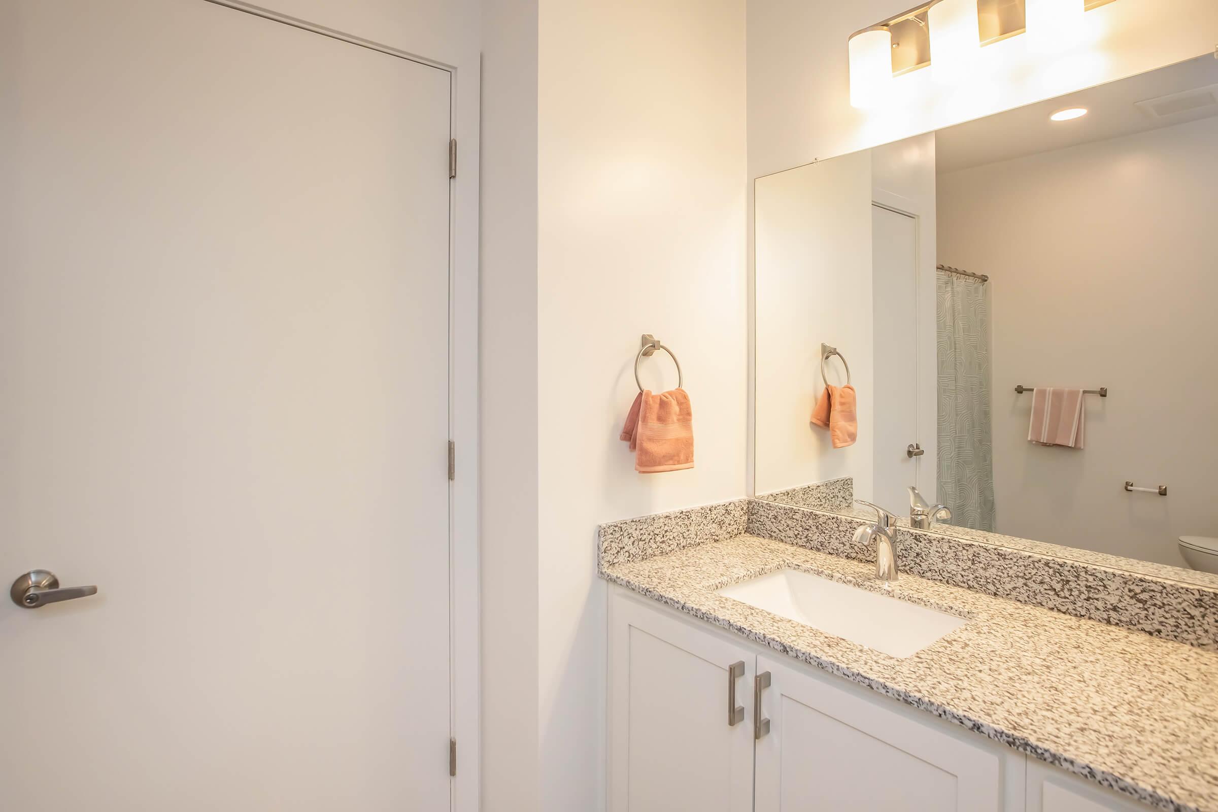 A modern bathroom featuring a light-colored wall with a mirrored cabinet above a granite countertop. The sink has a sleek faucet, and there are two hand towels hanging nearby. A shower curtain is visible in the background, and the doorway leads to another area. Soft lighting adds a warm ambiance.