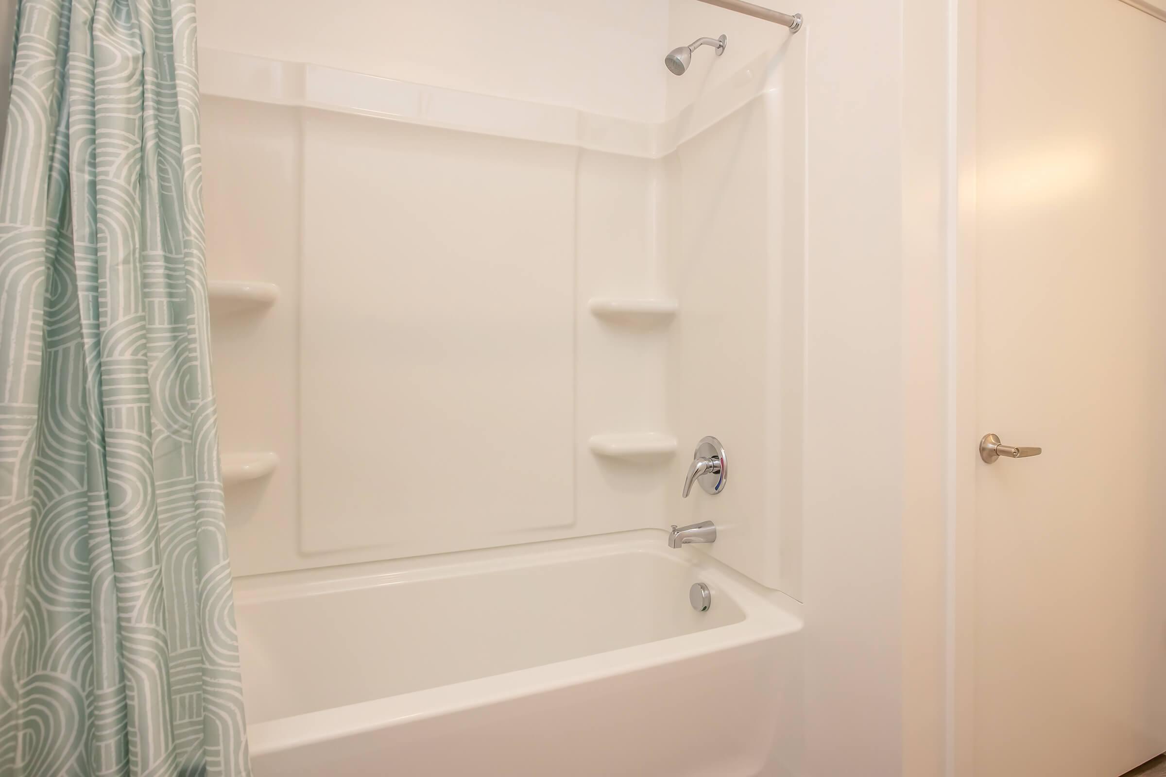A clean bathroom with a white bathtub, a light blue patterned shower curtain, and a showerhead. The walls are white, and there are two shelves built into the tub surround. A door is visible on the right side, completing the simple, minimalist design.