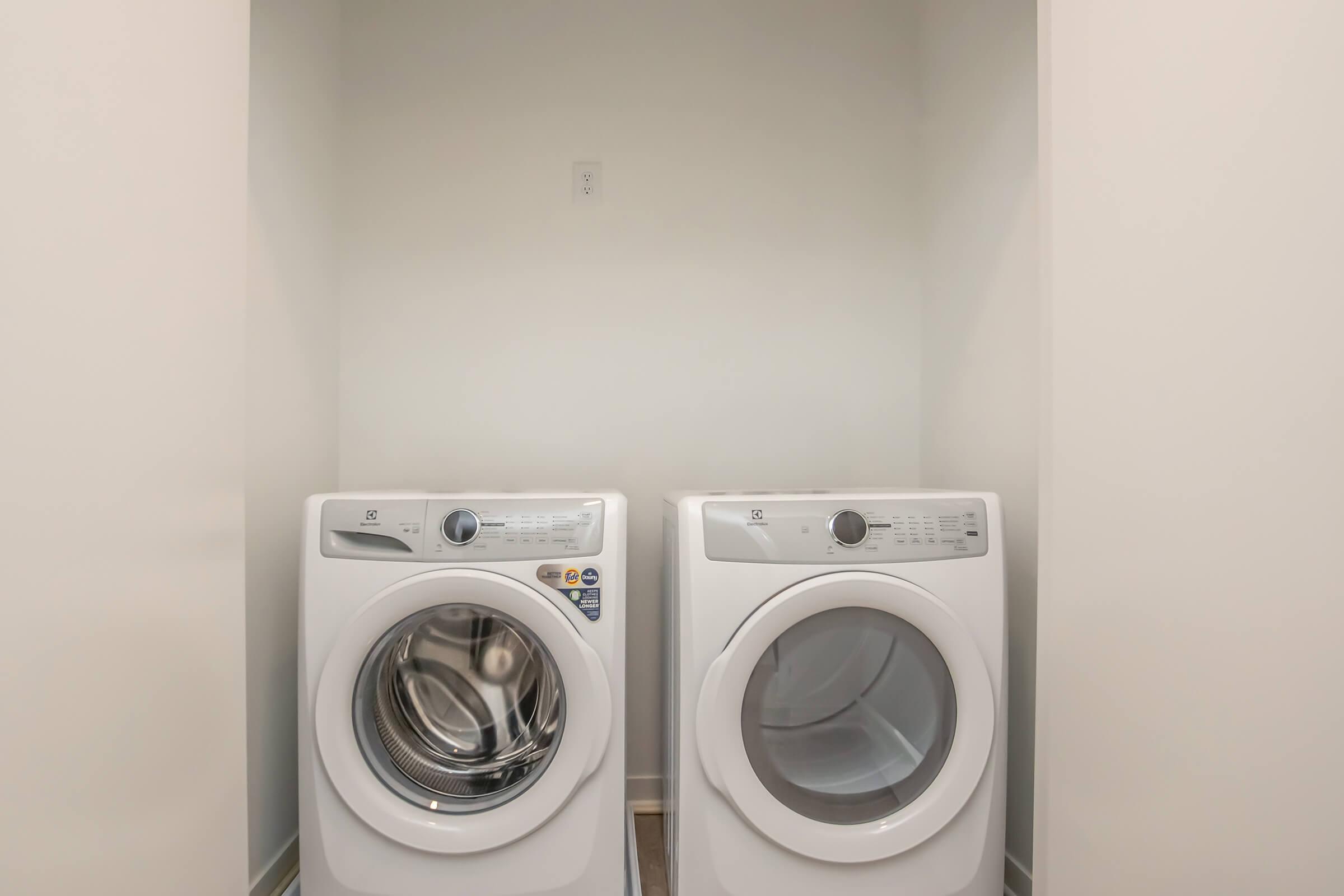 A laundry room featuring a pair of white washing machines side by side. The space is simple and minimalistic, with light-colored walls and a clean floor. There is a power outlet visible on the wall behind the machines.
