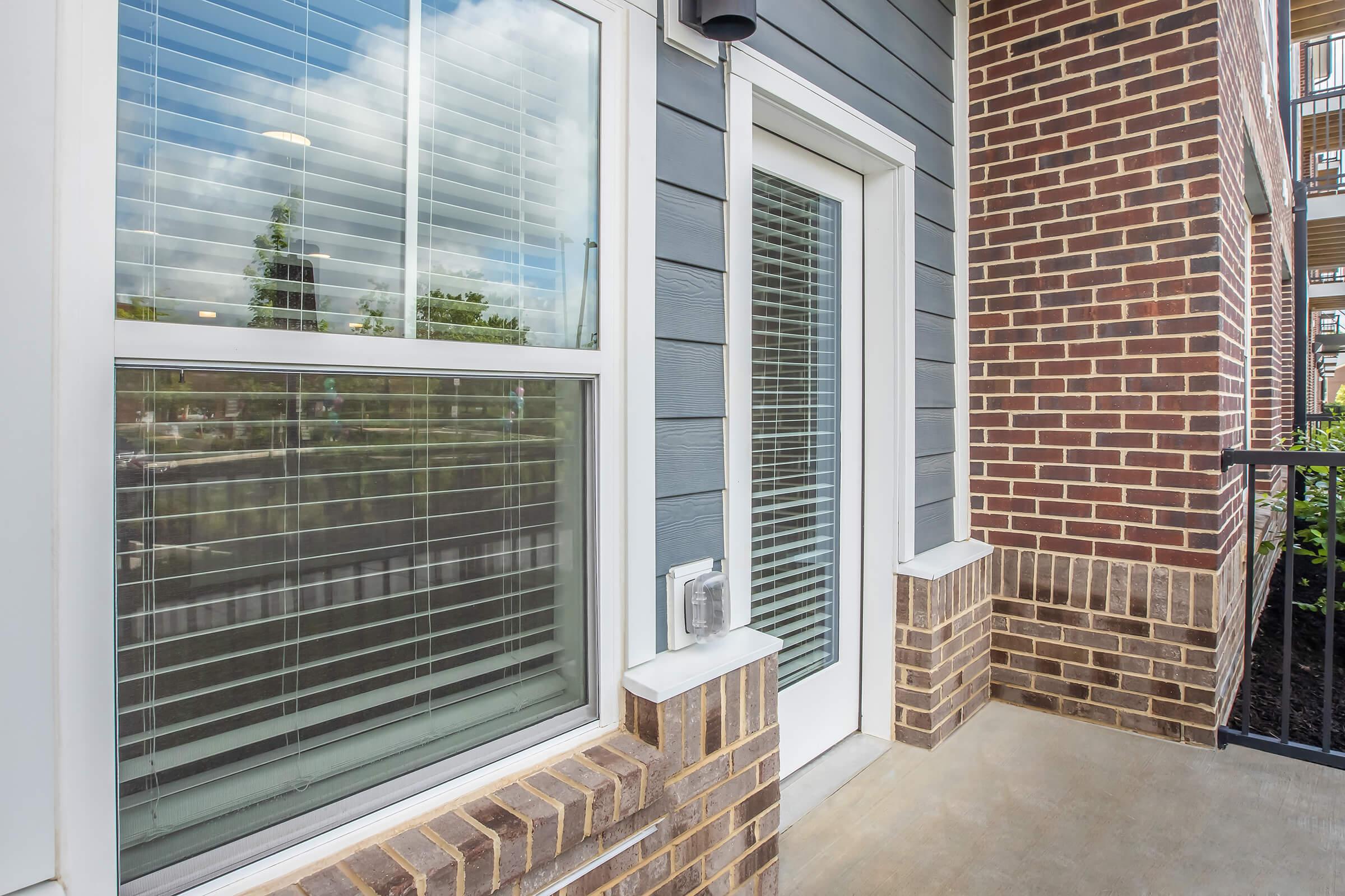 A close-up view of a modern balcony featuring a large window with blinds and a white door. The surrounding walls are made of smooth gray siding and textured brick. The balcony floor is concrete, and greenery is visible outside, with blue sky and clouds reflected in the window.