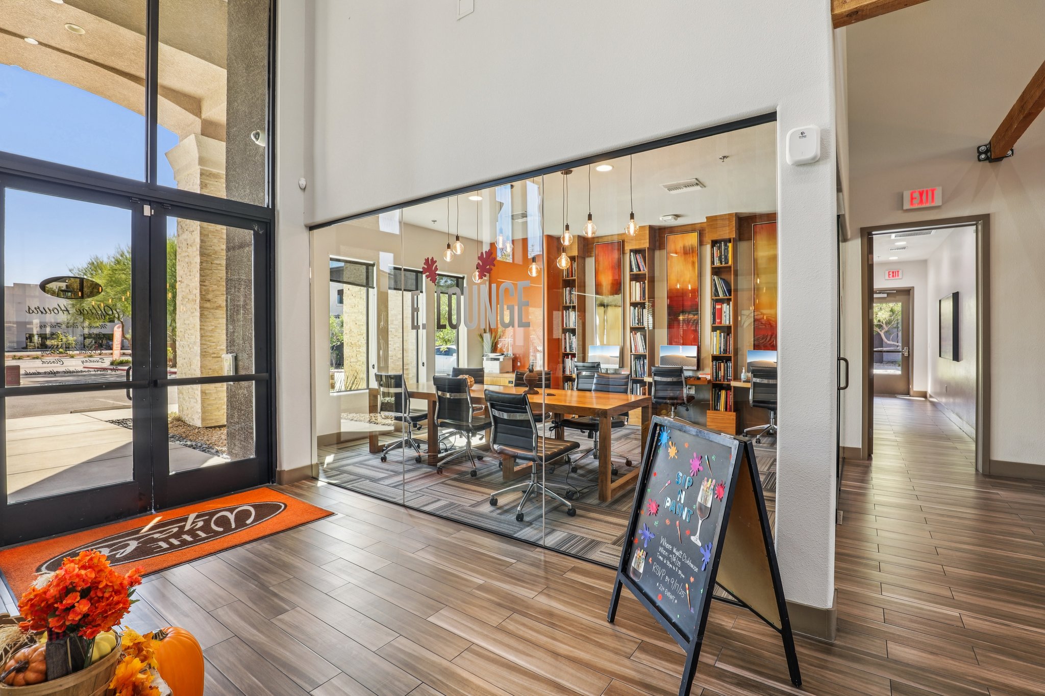 A modern lounge area featuring a large meeting table surrounded by ergonomic chairs, glass walls, and warm lighting. Decor includes decorative fall-themed elements, such as pumpkins. An entrance door is visible, leading to the outside. A chalkboard sign displays colorful art and messages.