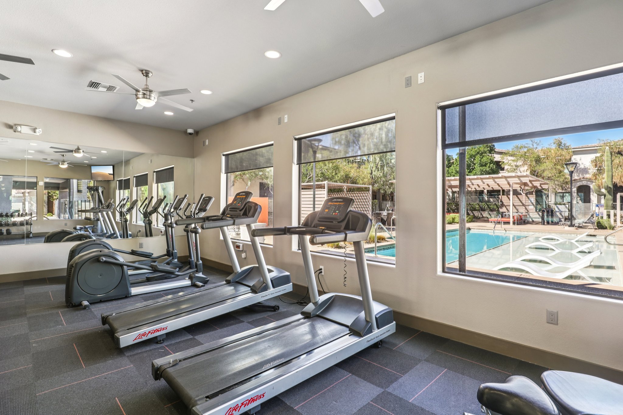 A modern gym interior featuring several treadmills lined up by large windows, showing a view of an outdoor swimming pool and lounge area. The space is well-lit with ceiling fans and mirrors, creating a bright and inviting atmosphere for exercise.