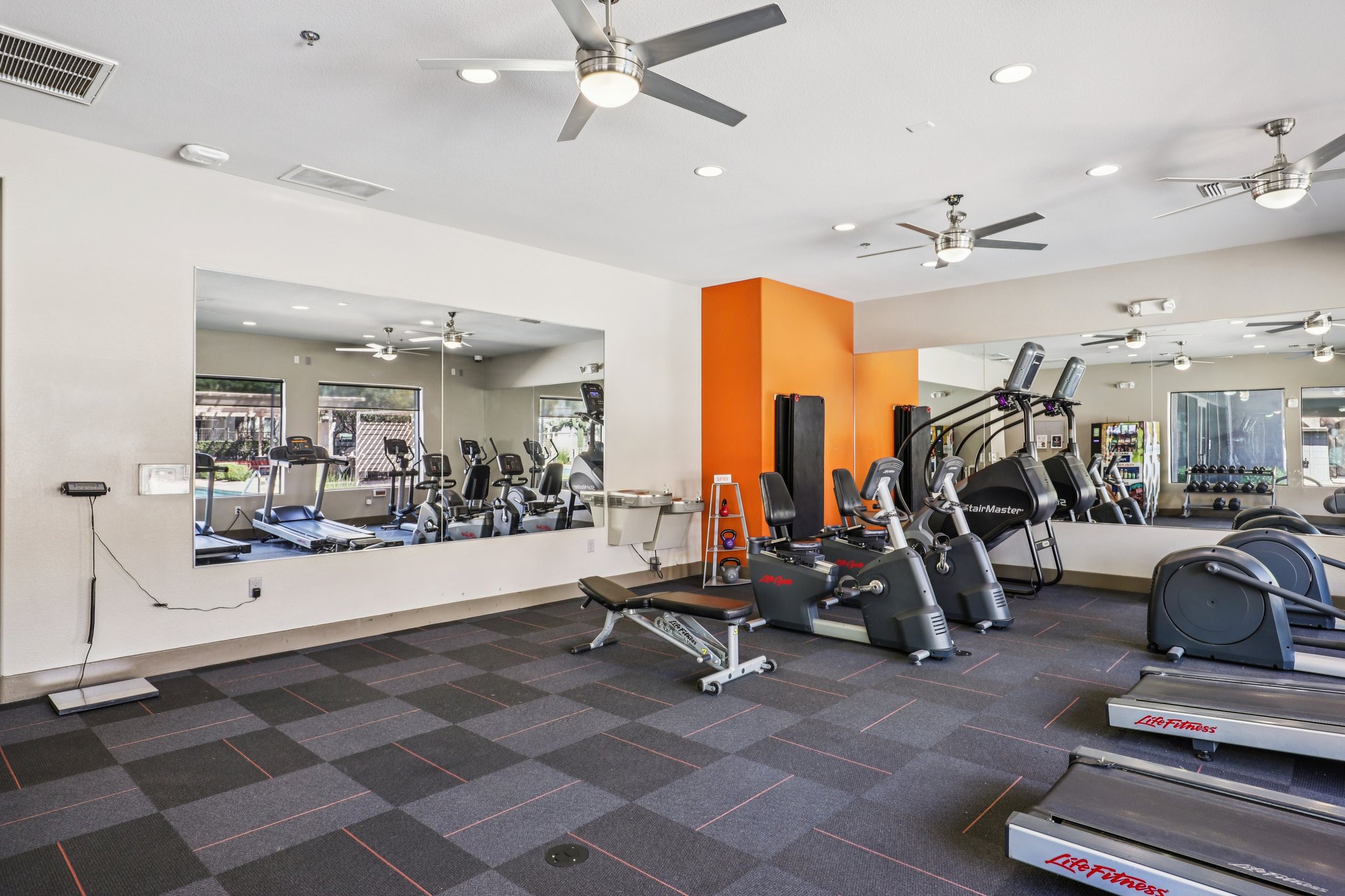 A well-lit gym area featuring exercise machines, including treadmills and ellipticals, along with free weights. Mirrors cover one wall, and there's a bright orange accent wall. The flooring consists of dark, patterned tiles, and ceiling fans are visible above for ventilation.
