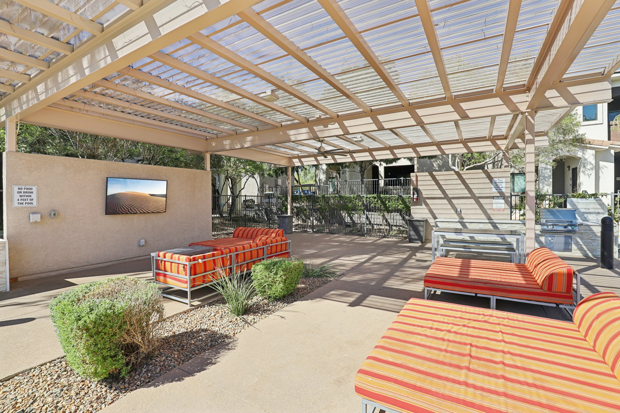 Outdoor lounge area with colorful striped seating, shaded by a pergola. A flat-screen TV is mounted on the wall, and there are small shrubs and gravel landscaping. The space is designed for relaxation and socializing in a residential setting.