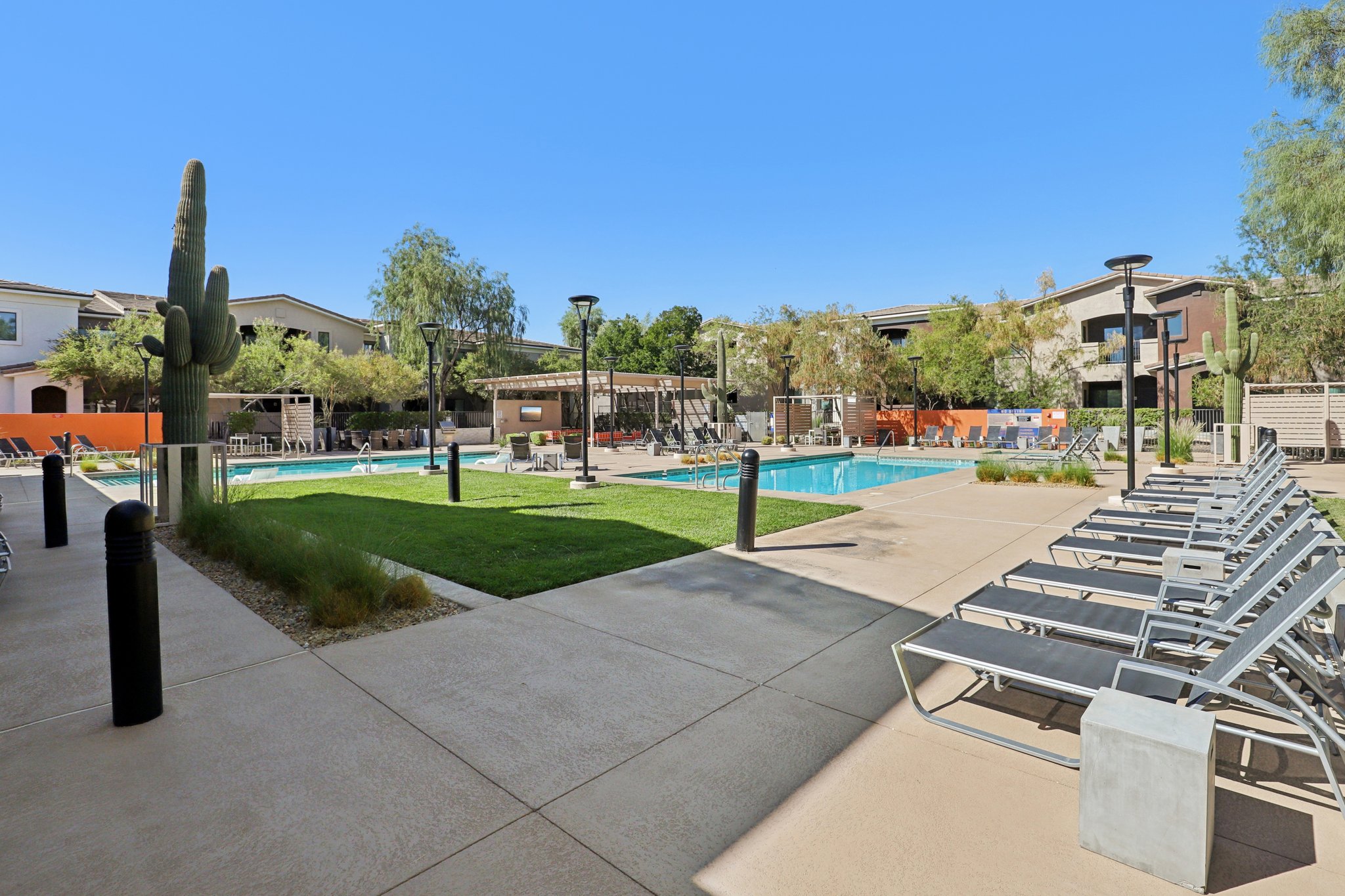 A sunny outdoor pool area featuring a swimming pool surrounded by lounge chairs, green grass, and decorative landscaping. Cacti and trees provide shade, and there are recreational facilities visible in the background. The clear blue sky adds to the inviting atmosphere.
