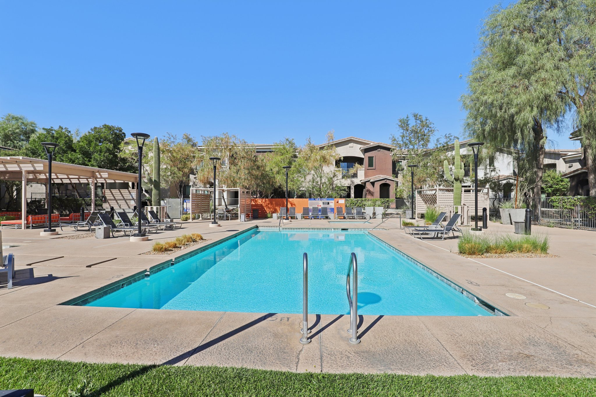 A clear swimming pool surrounded by palm trees and lounge chairs. There are shaded areas with seating, and apartment buildings are visible in the background under a bright blue sky. The pool area features clean, well-maintained concrete and green grass nearby.