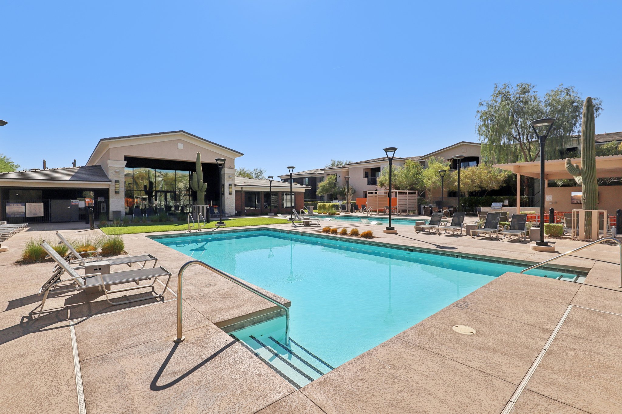 A spacious outdoor swimming pool surrounded by lounge chairs and lush greenery. Modern residential buildings are visible in the background, under a clear blue sky. The pool area is well-maintained, creating a relaxing atmosphere.