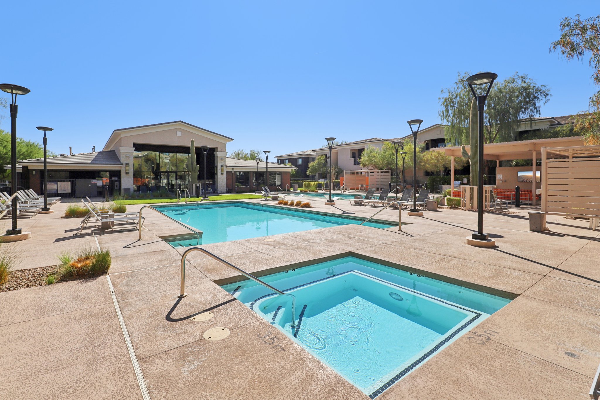 Two swimming pools in a sunny outdoor area, surrounded by lounge chairs and decorative plants. A hot tub is visible next to one of the pools. Buildings with entrances are seen in the background, along with palm trees and tall lamps, creating a resort-like atmosphere.