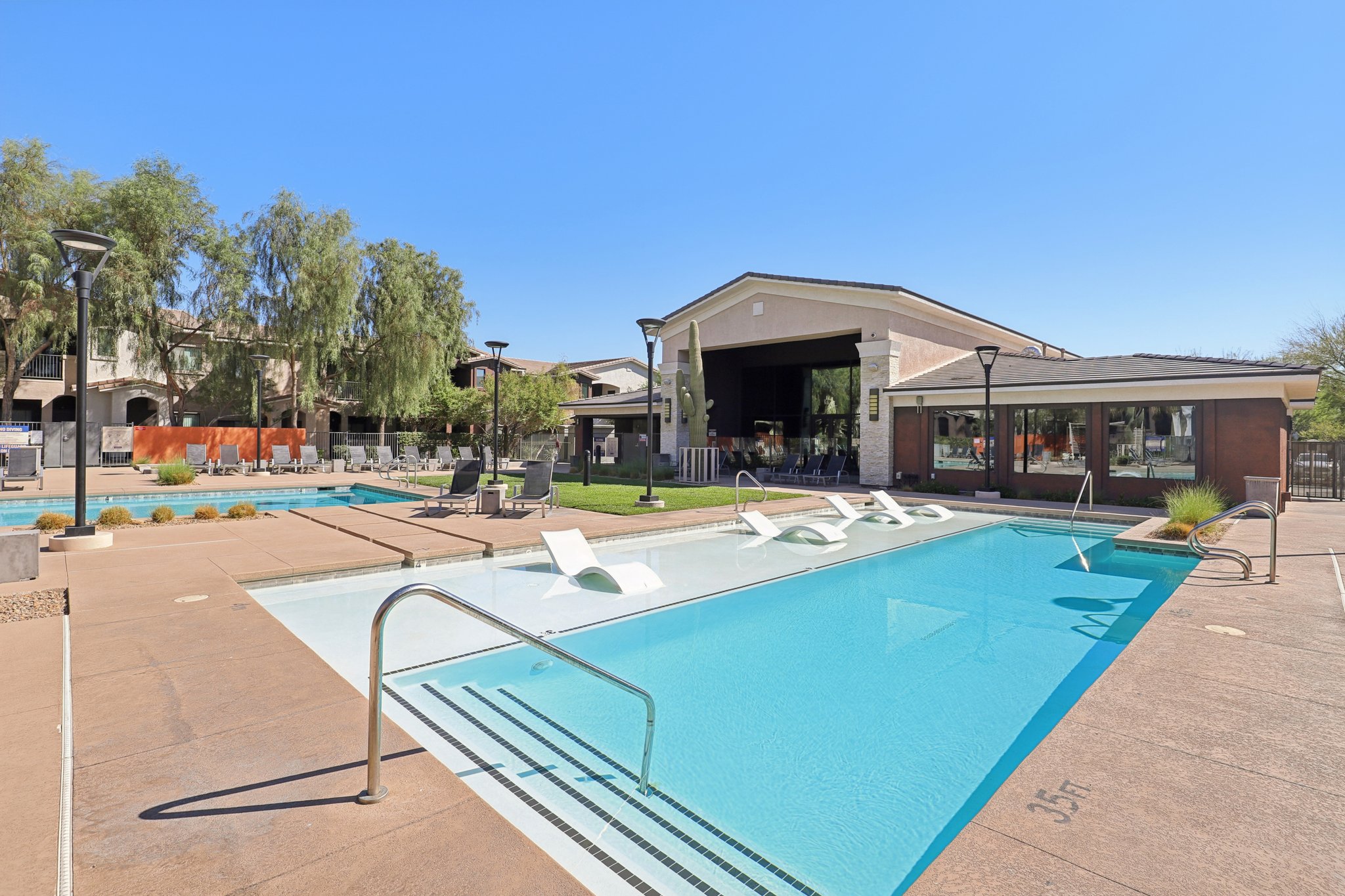 A bright outdoor swimming pool area featuring a spacious pool with lounge chairs, surrounded by landscaped gardens, and an adjoining building with large windows. The sky is clear blue, creating a relaxing atmosphere for recreation and leisure.