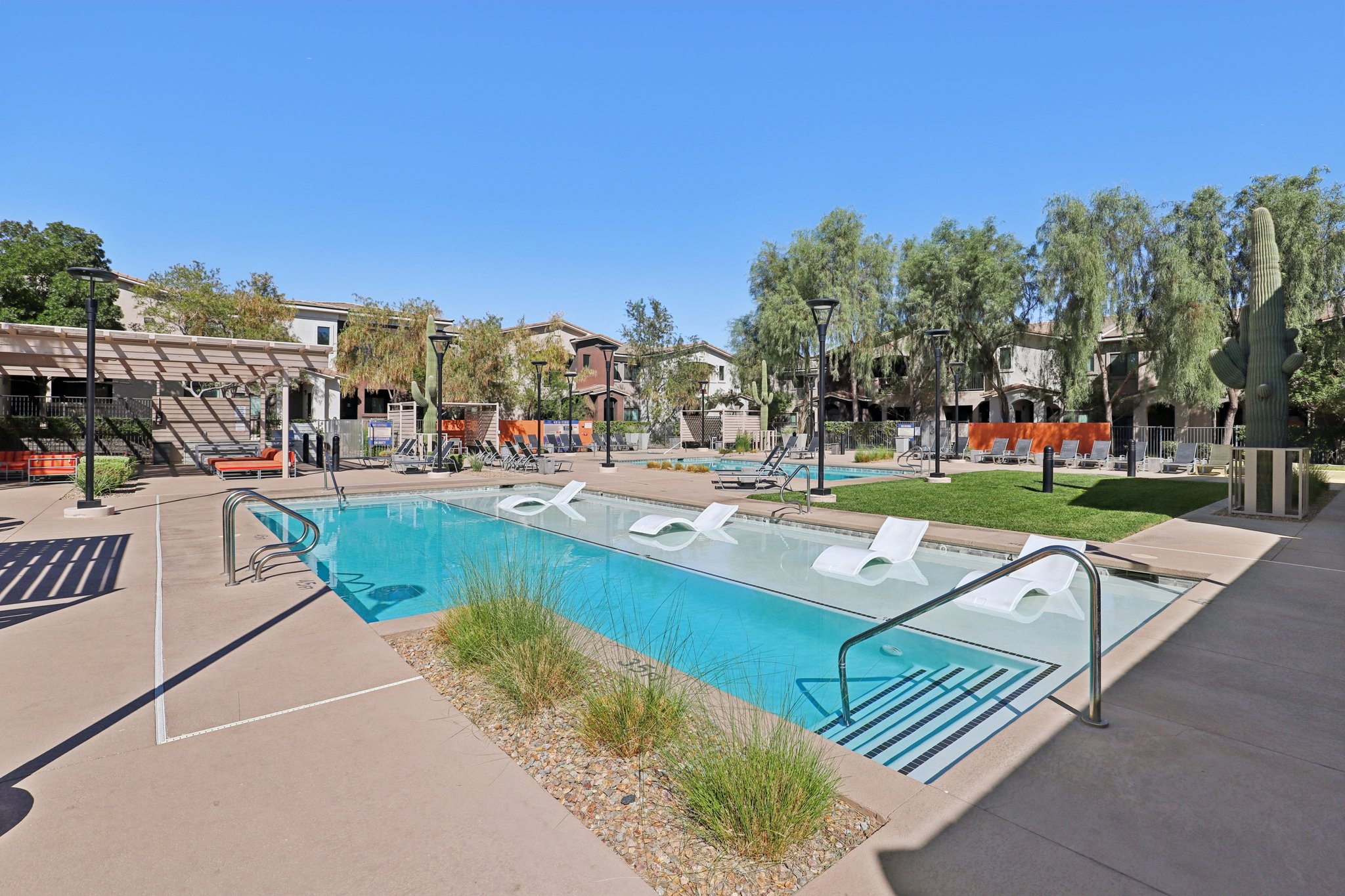 A modern outdoor swimming pool area surrounded by lounge chairs and green landscaping. In the background, there are residential buildings and shaded seating areas under pergolas. The clear blue sky enhances the inviting atmosphere of the space, ideal for relaxation and social activities.