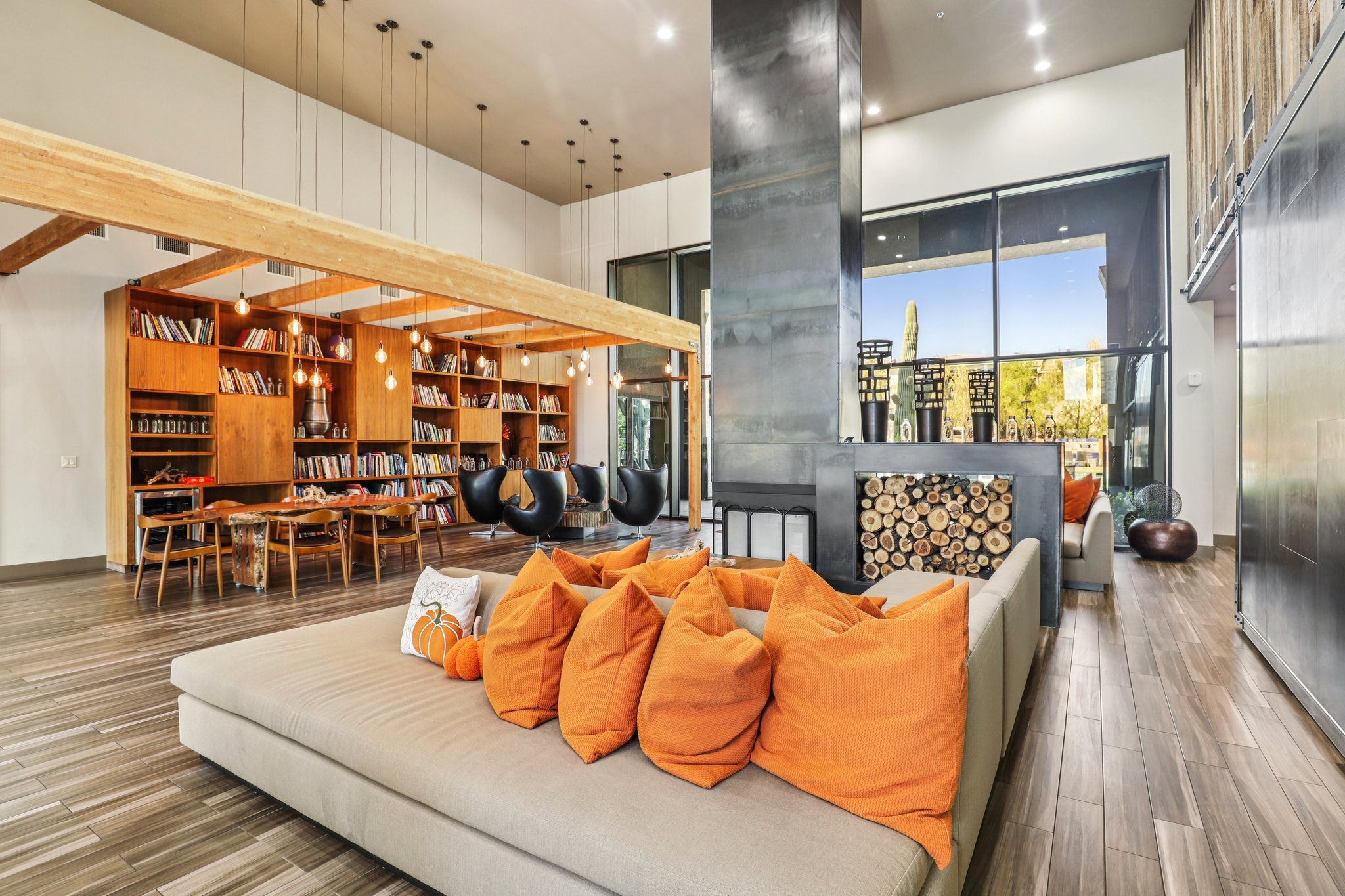 A modern lounge area featuring a cozy sectional sofa adorned with orange decorative pillows. The space includes large windows, wooden shelving filled with books, a dining table with chairs, and unique decor elements. Soft lighting from hanging fixtures creates a warm and inviting atmosphere.