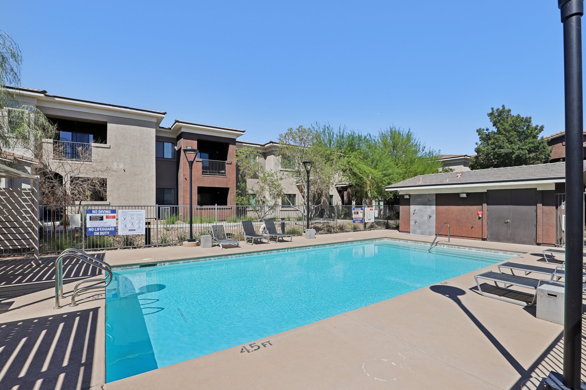 A clean, inviting swimming pool surrounded by lounge chairs, set in a landscaped area with residential buildings in the background. The sky is clear and blue, and there are signs near the pool indicating rules and information.
