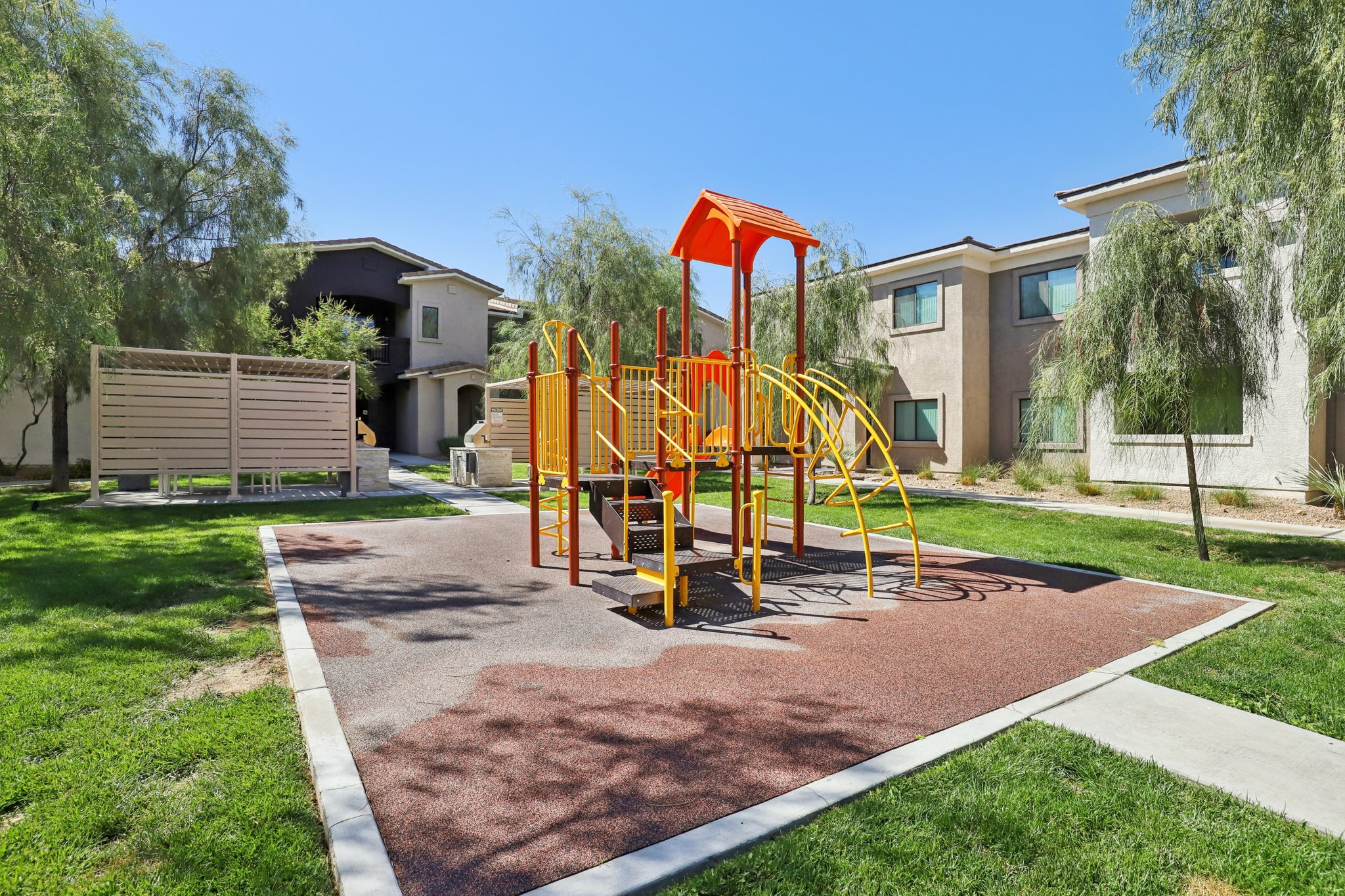 A colorful playground with orange and yellow equipment is set on a rubber surface in a grassy area. Surrounding buildings are visible in the background, along with some trees providing shade. The sky is clear and blue, indicating a sunny day.