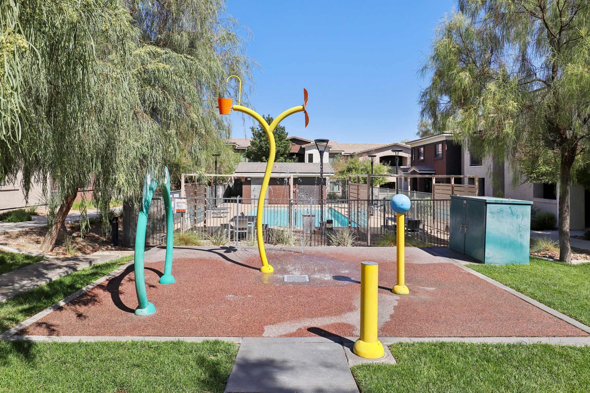 A colorful water play area in a residential setting features three whimsical, tall sprouting water fixtures: one with a bucket, another with a spray nozzle, and a third with a rotating ball. Surrounding the area are green grass and trees, with a fenced pool visible in the background under a clear blue sky.