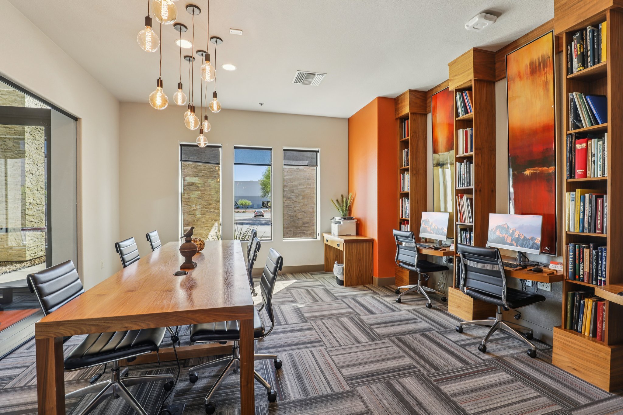 A modern meeting room featuring a long wooden table surrounded by black chairs, with two computer stations against a wall of bookshelves filled with books. Large windows let in natural light, and an accent wall painted in orange adds warmth to the space. Stylish pendant lights hang from the ceiling.