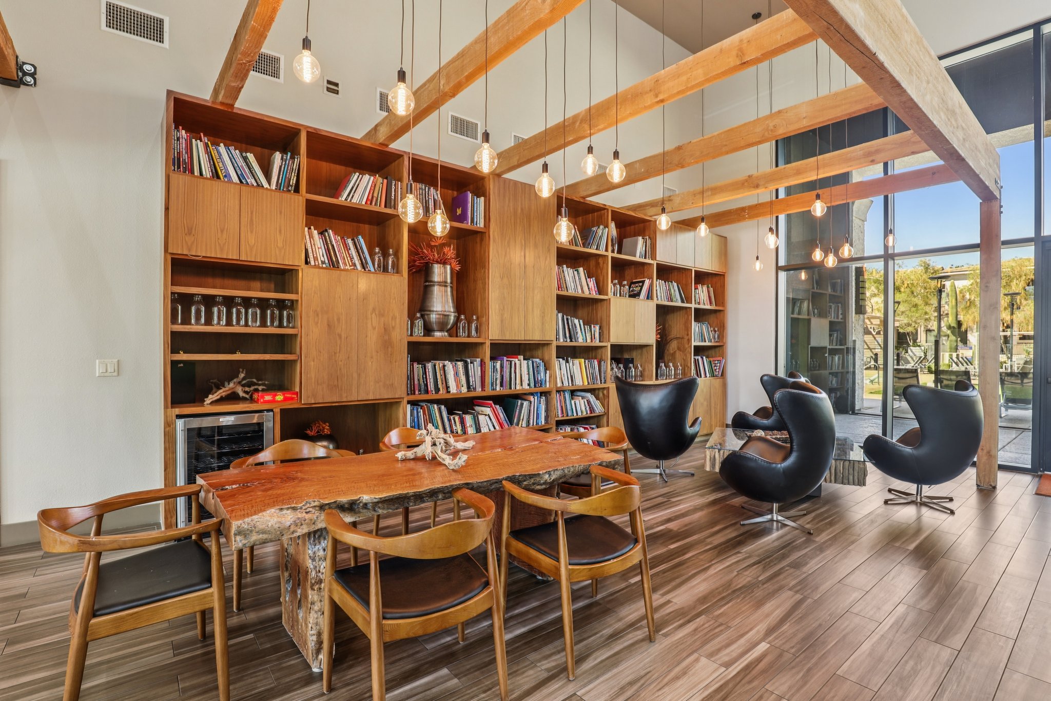Cozy interior of a modern reading room featuring a wooden bookshelf filled with books, a dining table surrounded by wooden chairs, and stylish black lounge chairs. The space is illuminated by pendant lights and has large windows allowing natural light. Elements of nature are incorporated into the decor.