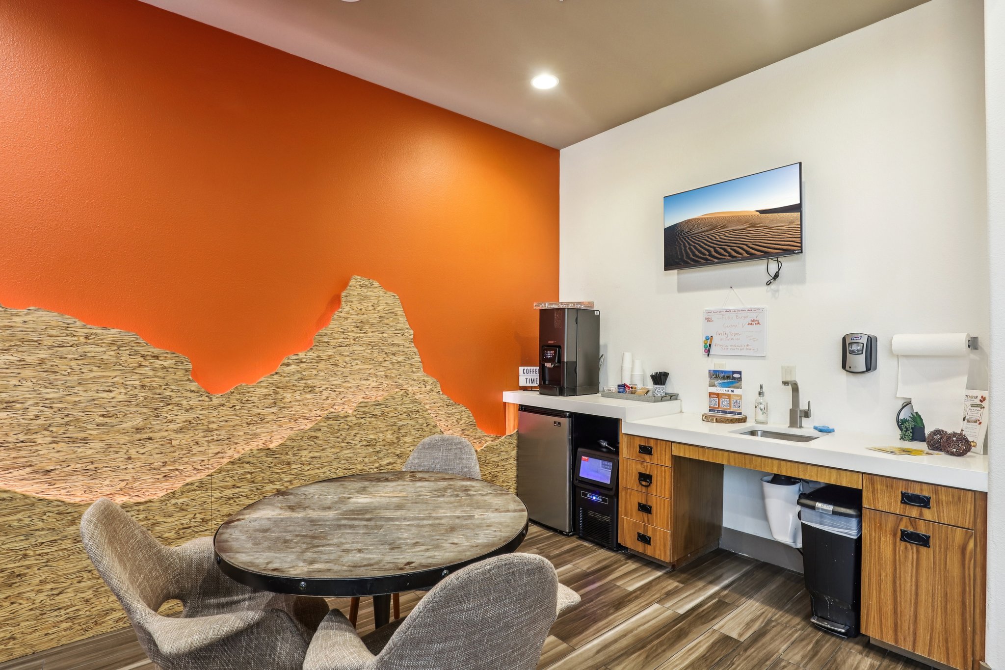 A modern kitchenette featuring an orange accent wall and a textured wall design. It includes a round wooden table with gray chairs, a mini fridge, a countertop with a sink, and a wall-mounted TV displaying a desert landscape. The area appears well-lit and inviting.