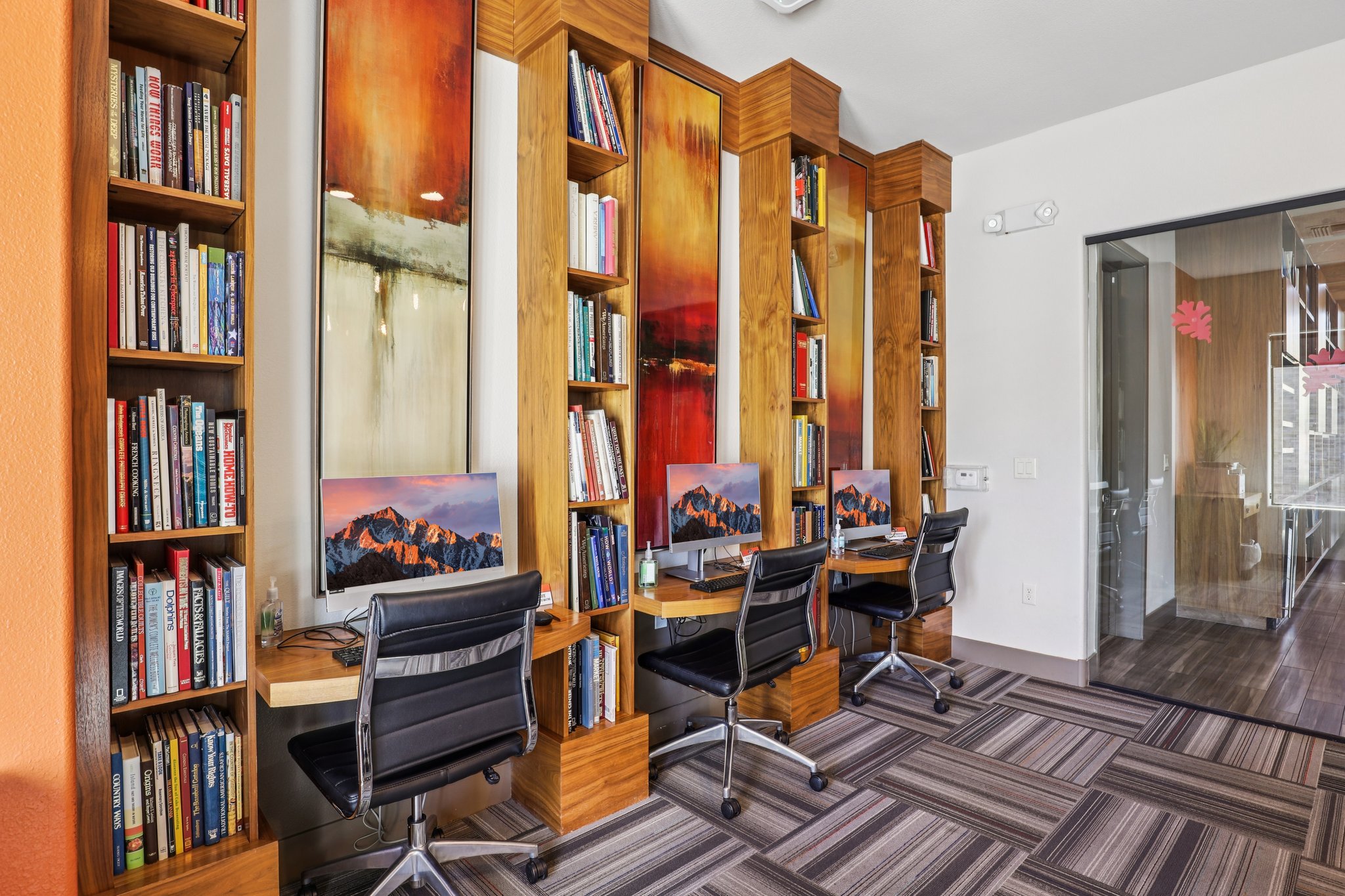 A modern, stylish library or study area featuring tall wooden bookshelves filled with books, two computer workstations with monitors, and contemporary decor including colorful wall art. The space has a light and inviting atmosphere, with a mix of natural and artificial lighting.
