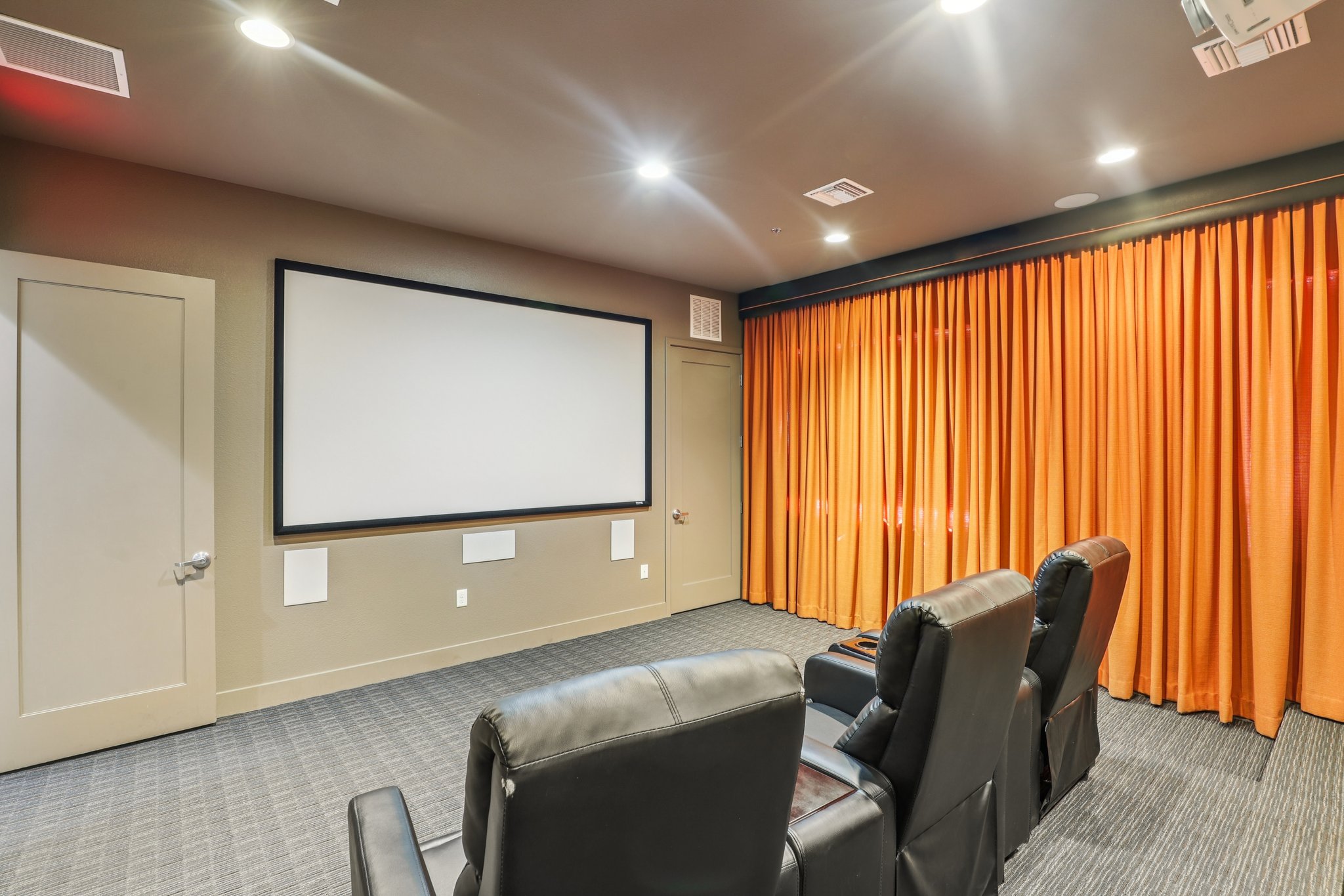 A cozy home theater room featuring a large blank projection screen on the wall, three comfortable black recliners facing the screen, and vibrant orange curtains. The room is well-lit with recessed ceiling lights, creating a welcoming ambiance for movie watching.