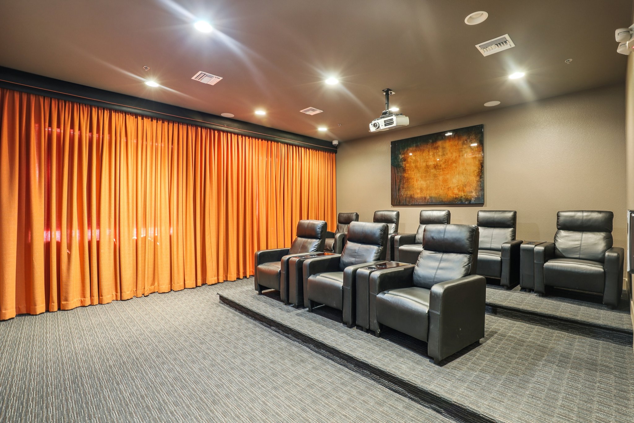 Home theater with black reclining chairs arranged in tiers. The walls are painted in a neutral tone, and there's an overhead projector mounted. Bright orange curtains cover the windows, and a large artwork hangs on the wall. The room is well-lit with ceiling lights.