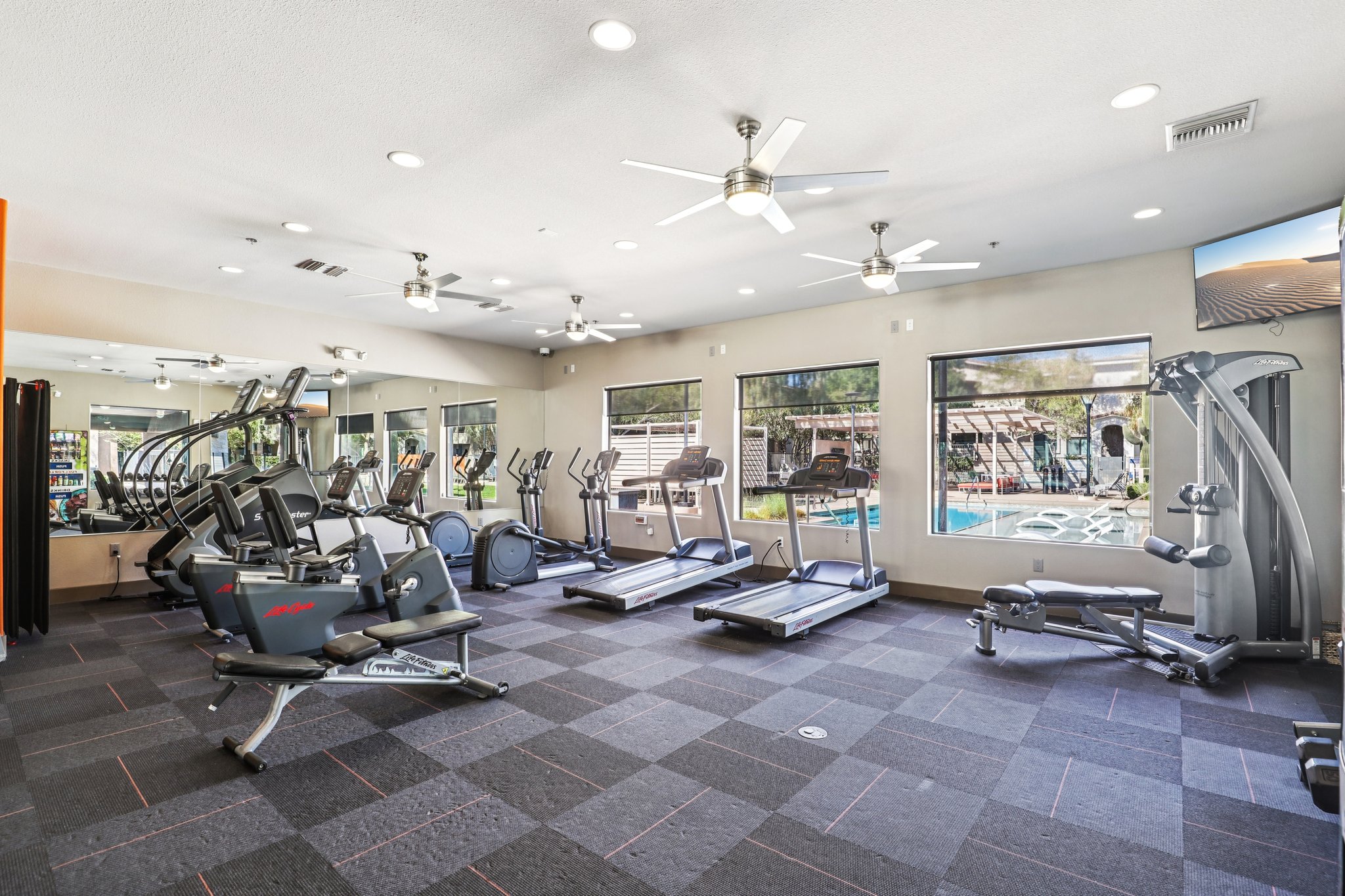 A modern gym with various exercise equipment including treadmills, ellipticals, and strength training machines. The room features large windows with views of a pool area outside, bright overhead lights, and ceiling fans. The flooring is dark with a textured pattern, creating a spacious and well-lit workout environment.