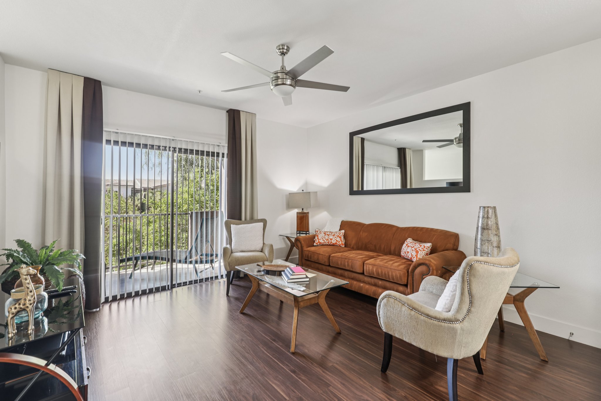 Interior of a modern living room featuring a brown leather sofa, two cream-colored armchairs, and a glass coffee table. A large mirror hangs on the wall opposite the sofa. Natural light fills the space through sliding glass doors leading to a balcony, framed by long curtains. Decorative plants and items are arranged around the room.