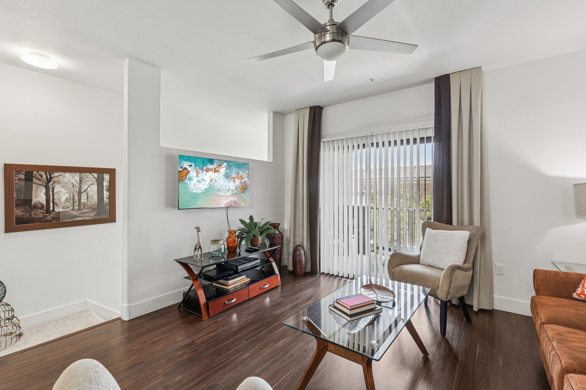 Modern living room featuring a ceiling fan, large windows with vertical blinds, and a glass coffee table. One wall is adorned with two artworks, while a stylish TV stand displays decorative items. A comfortable chair and a sofa add to the cozy atmosphere, with natural light enhancing the space.