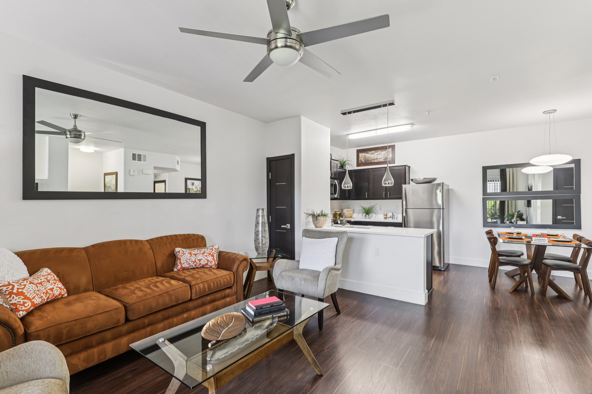 Modern open-concept living area featuring a brown sofa with decorative pillows, a glass coffee table, and a dining area with wooden chairs. The kitchen is equipped with stainless steel appliances, and there are stylish light fixtures hanging above. A large mirror adds depth to the space, enhancing the contemporary aesthetic.