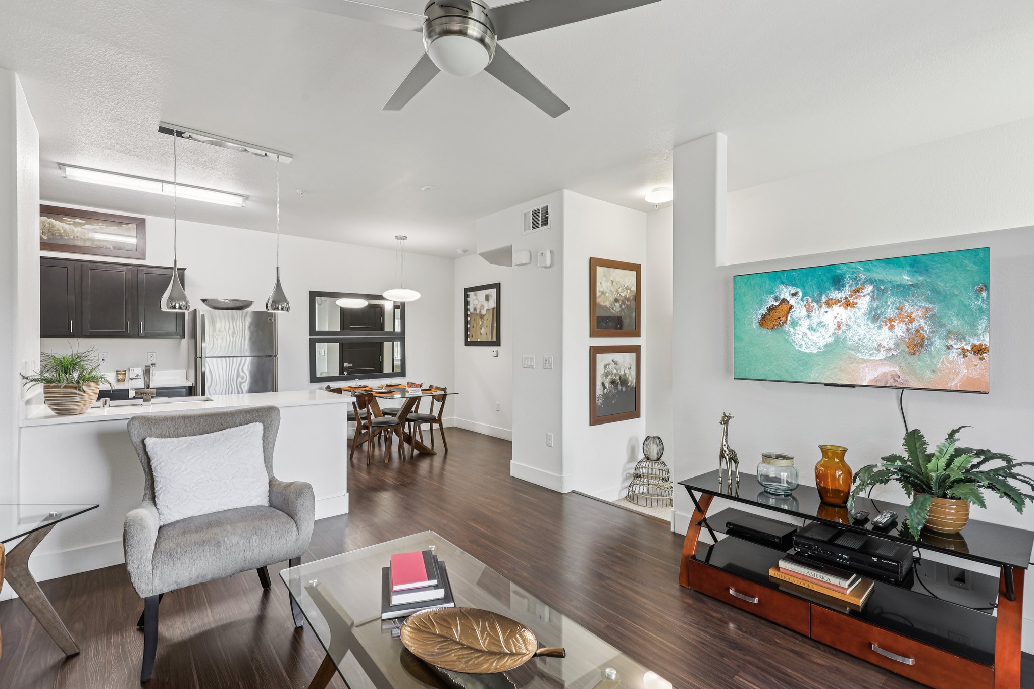 A modern living room featuring a gray armchair, glass coffee table, wooden floor, and a flat-screen TV mounted on the wall displaying a beach scene. In the background, there's a dining area with a wooden table and chairs, and a kitchen with dark cabinetry and stainless-steel appliances.