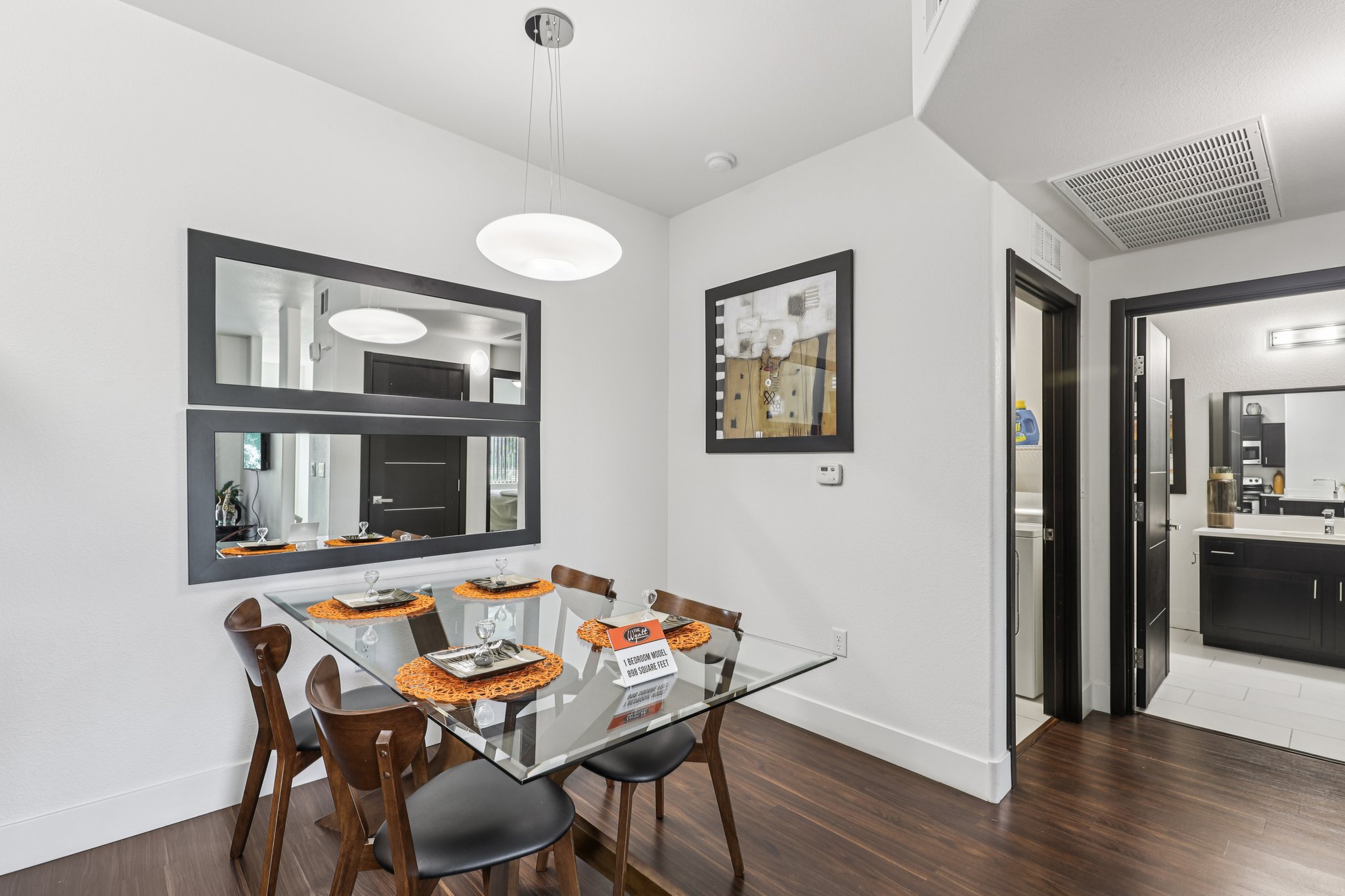 A modern dining area featuring a glass table set with orange placemats and plates, surrounded by wooden chairs. The walls are painted white, with a large mirror and framed artwork. An entrance leads to a bathroom, and a sleek ceiling light illuminates the space.