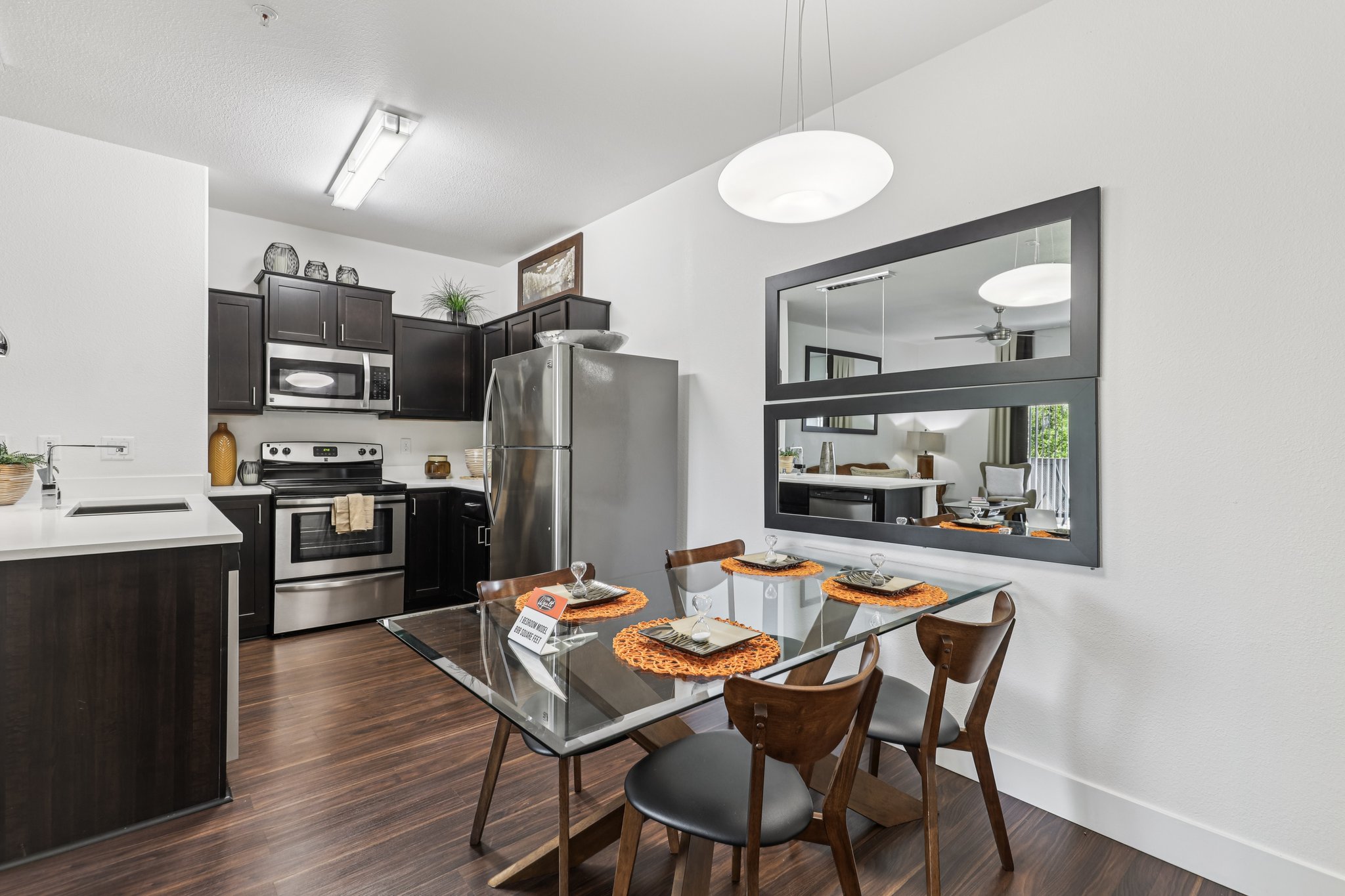 Modern kitchen and dining area featuring a sleek stainless steel refrigerator, an oven, and dark wooden cabinetry. A glass dining table is set with plates and orange placemats, accompanied by wooden chairs. A decorative mirror reflects light and adds depth to the space, with a cozy living area visible in the background.