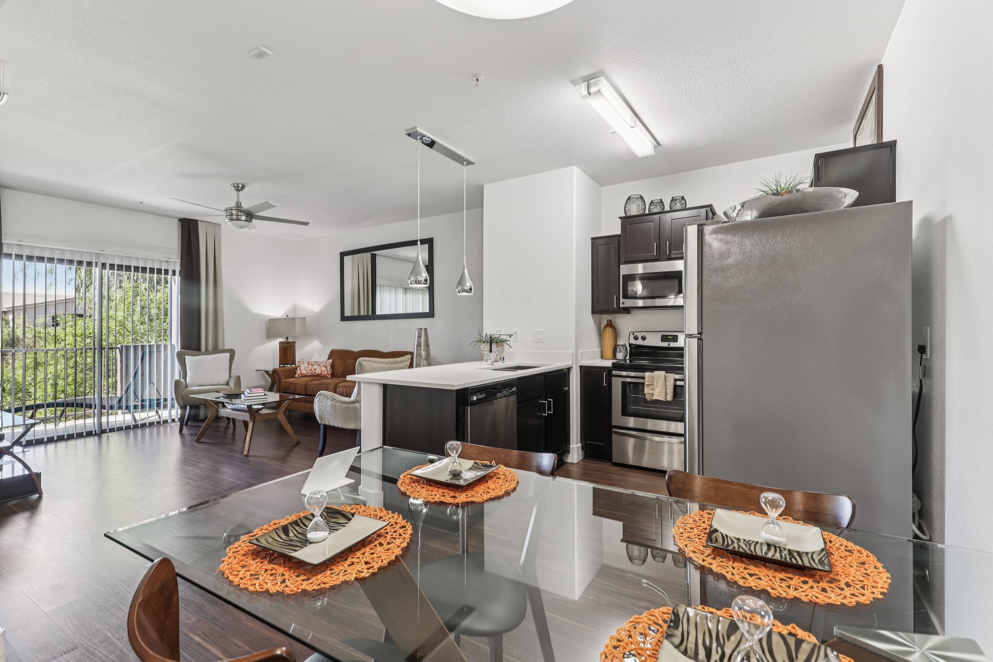 Modern open-plan kitchen and dining area featuring a glass dining table set for four with orange placemats. The kitchen showcases dark cabinetry, stainless steel appliances, and a large island. A comfortable living area is visible in the background, complete with a sofa and sliding glass doors leading to an outdoor space.