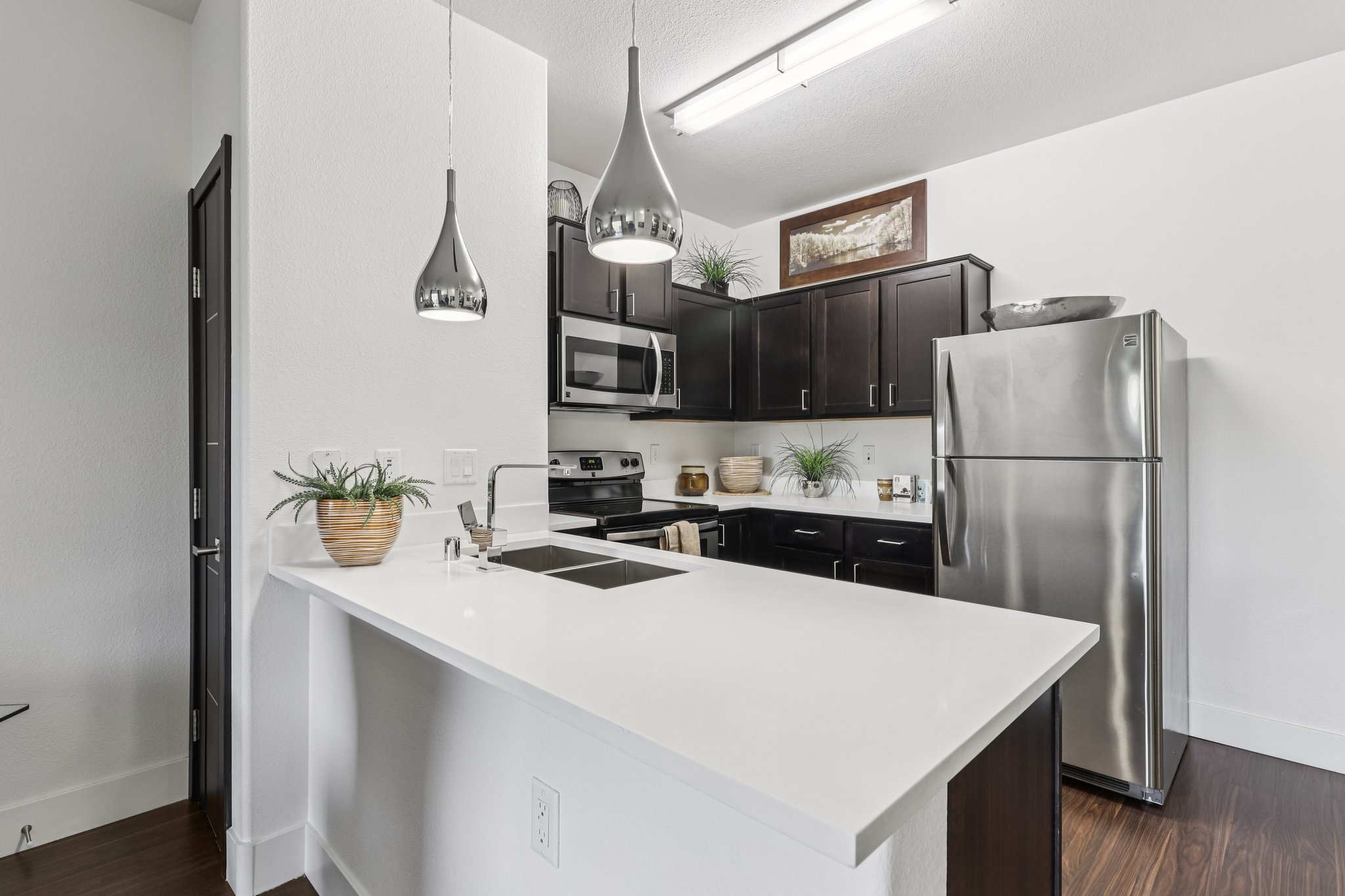Modern kitchen featuring dark cabinets, stainless steel appliances, and a white countertop. The space includes a sink on the island, decorative plants, and pendant lighting for a sleek, contemporary look. The overall design is open and bright, with a minimalist aesthetic.