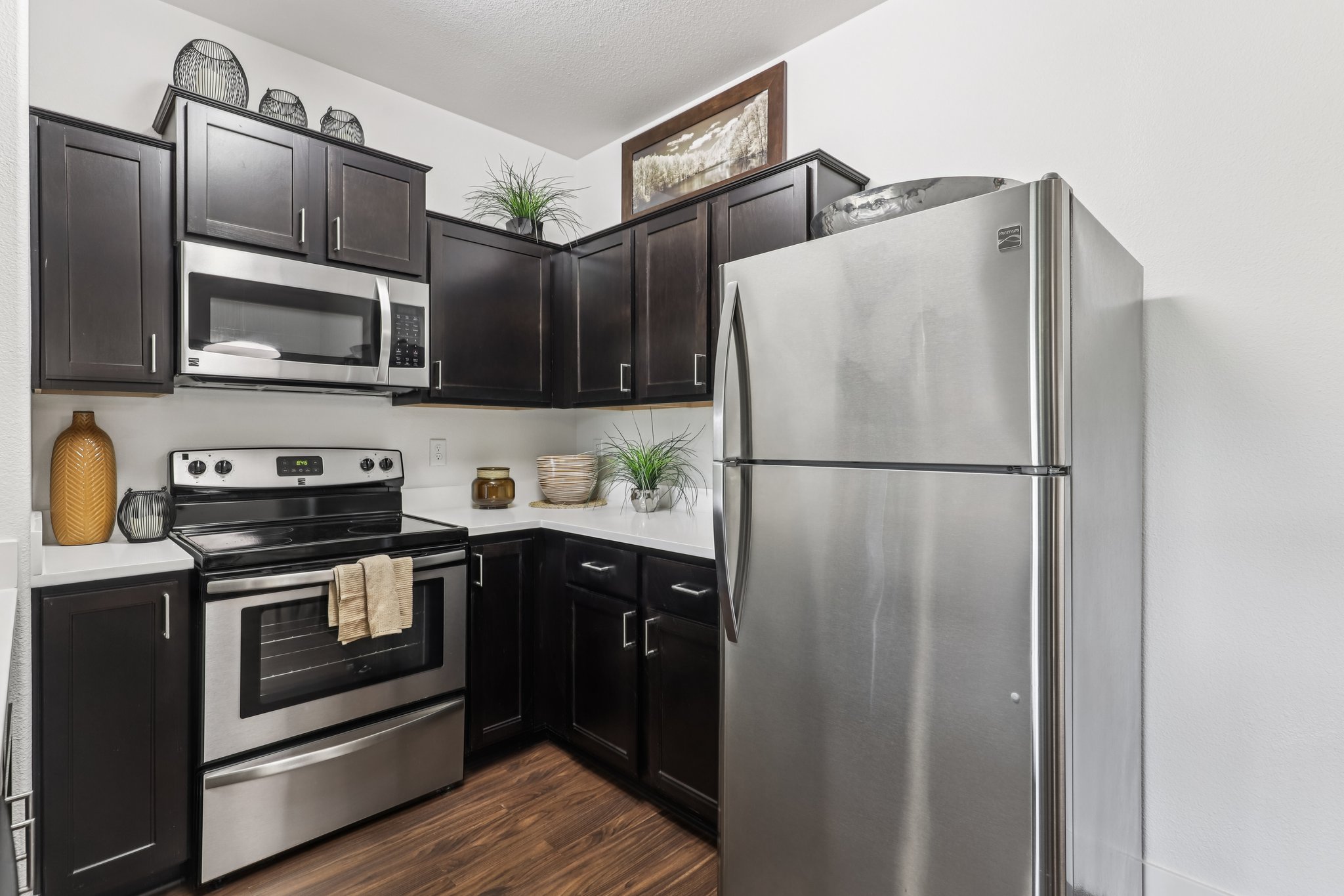A modern kitchen featuring dark wood cabinets, stainless steel appliances, and a sleek countertop. It includes a microwave, stovetop oven, and a large silver refrigerator. Decorative items like plants and bowls are arranged on the shelves to add a touch of warmth and style.