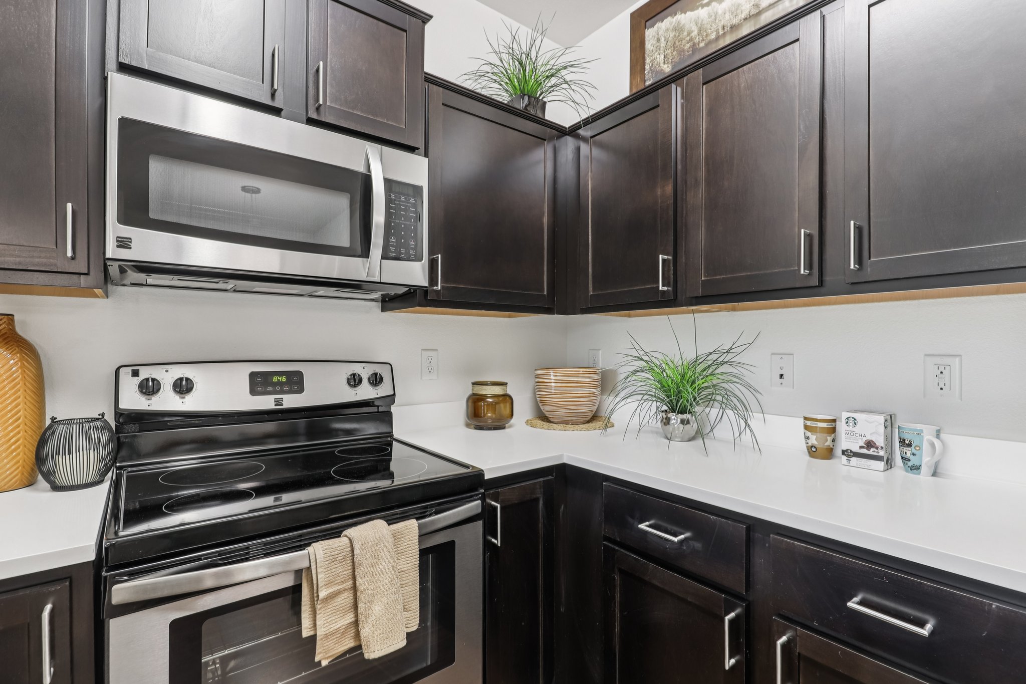 A modern kitchen featuring dark wooden cabinets, a stainless steel microwave and oven, and a white countertop. Decorative items include a woven basket, small plants, and jars on the counter, creating a stylish and inviting atmosphere.