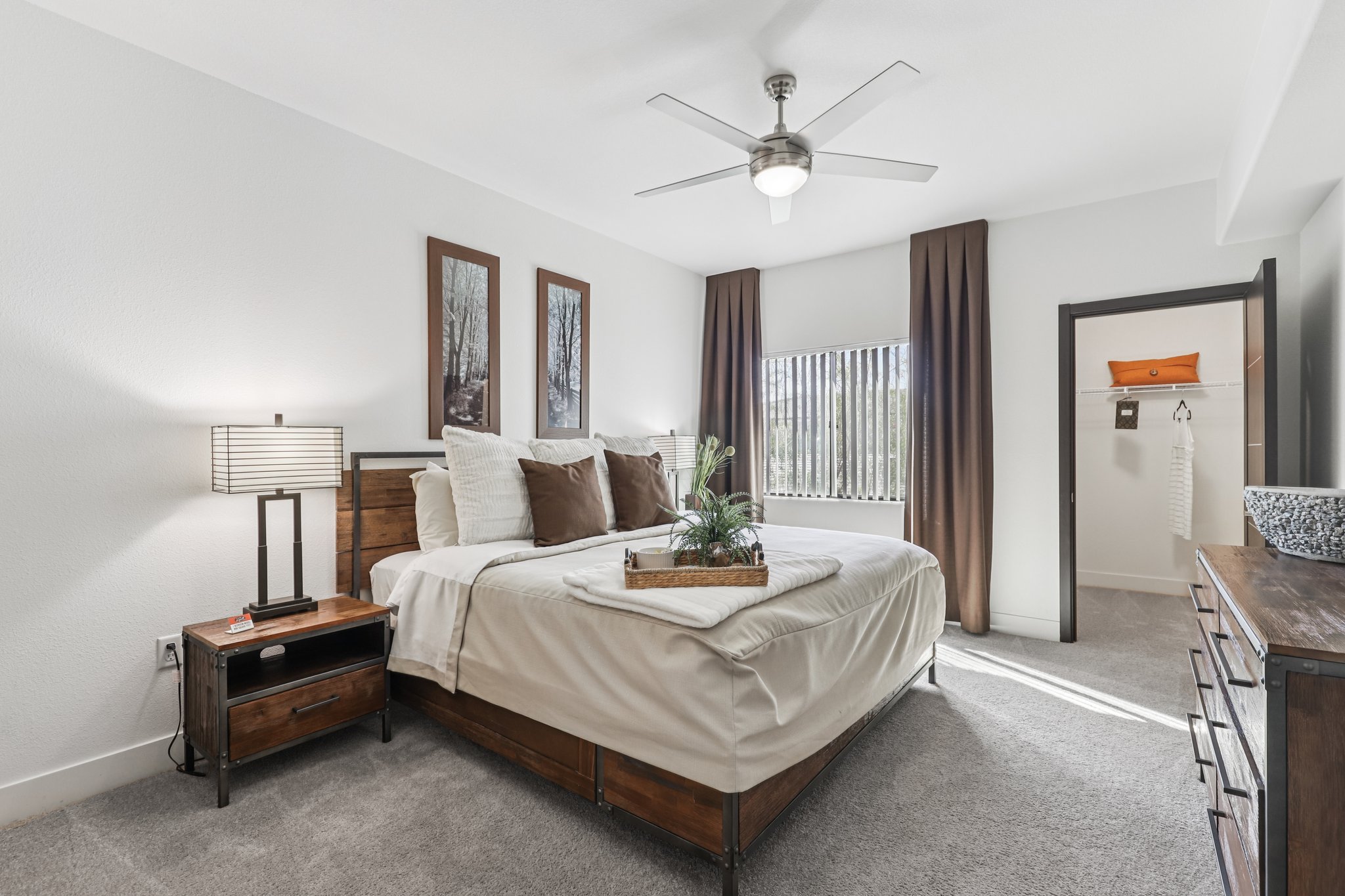 A modern bedroom featuring a neatly made bed with decorative pillows, a nightstand with a lamp, and a ceiling fan. Two framed artworks hang above the bed. Natural light filters through the tall window with curtains, and there is a closet visible in the background. The decor has a warm, inviting ambiance.