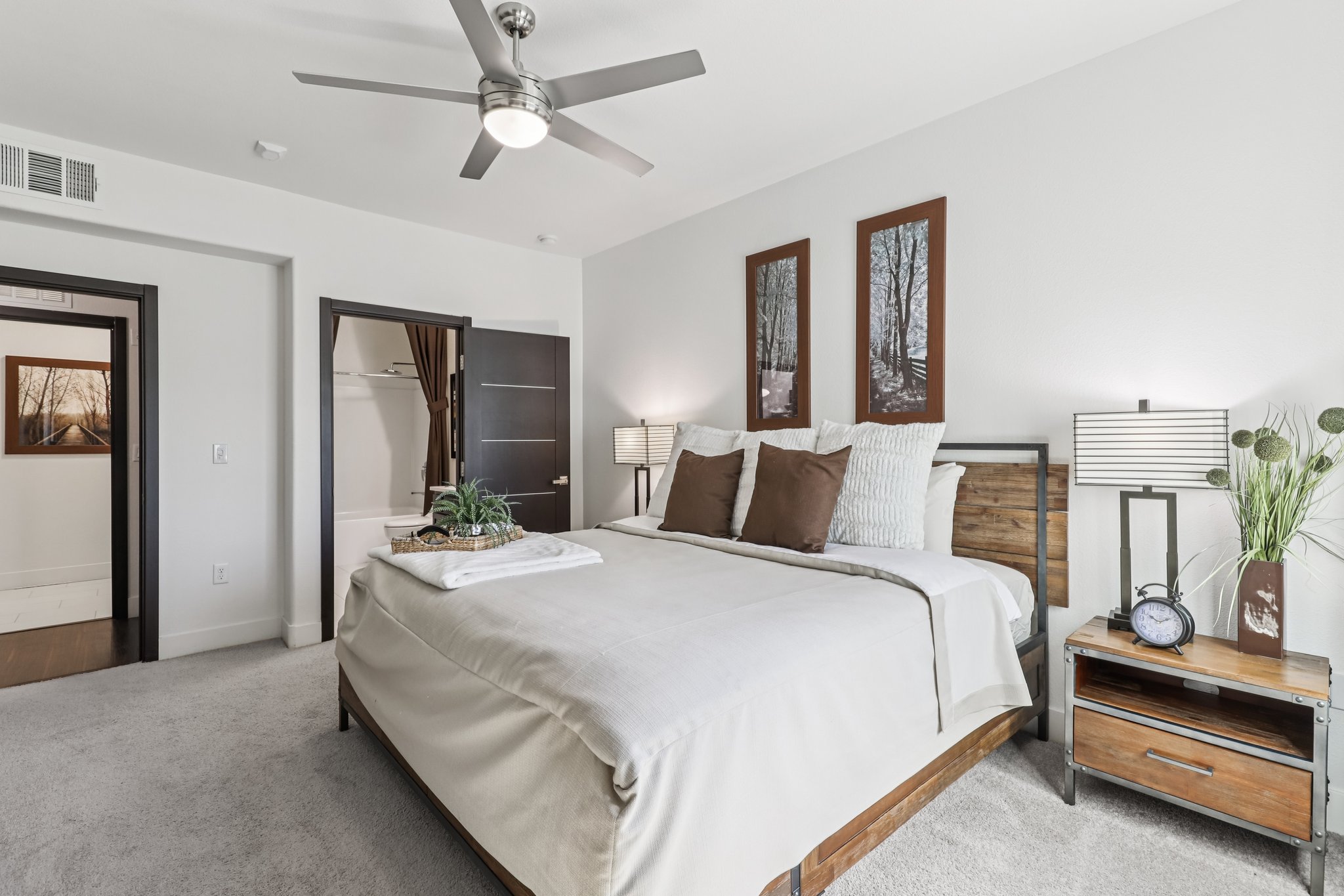 A modern bedroom featuring a queen-sized bed with a light-colored blanket and decorative pillows. There are two bedside lamps on wooden nightstands, a clock, and a small plant. The walls are painted white, and there are artwork frames above the bed. A door leads to a bathroom area, and the floor is carpeted.
