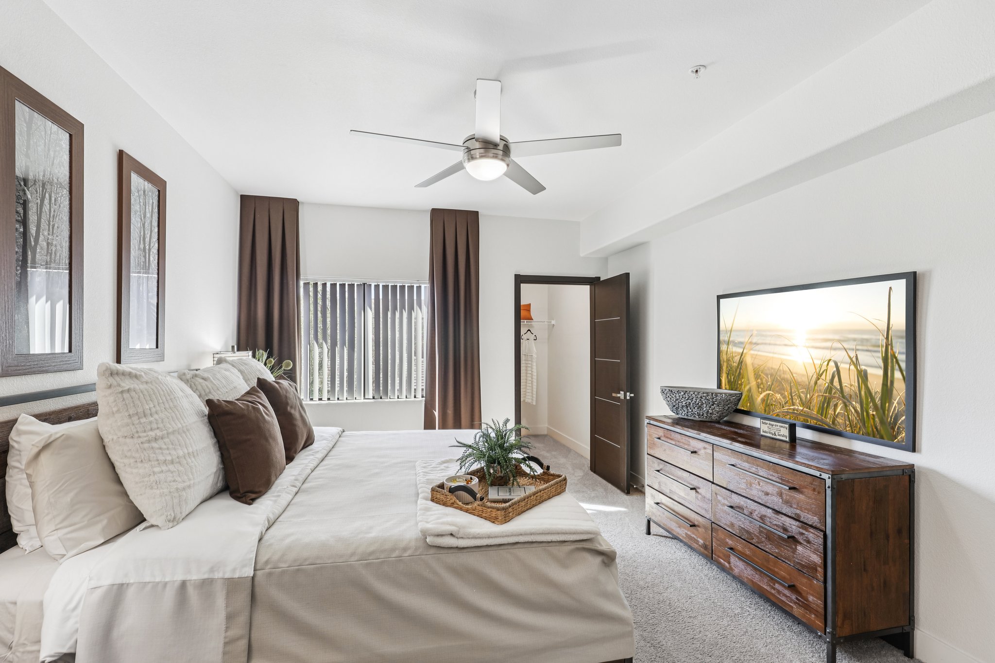 A modern, well-decorated bedroom featuring a comfortable bed with decorative pillows, a wooden dresser, a large TV displaying a serene landscape, and a ceiling fan. Natural light filters through blinds, with stylish wall art and curtains enhancing the aesthetic appeal. A cozy atmosphere with neutral tones.