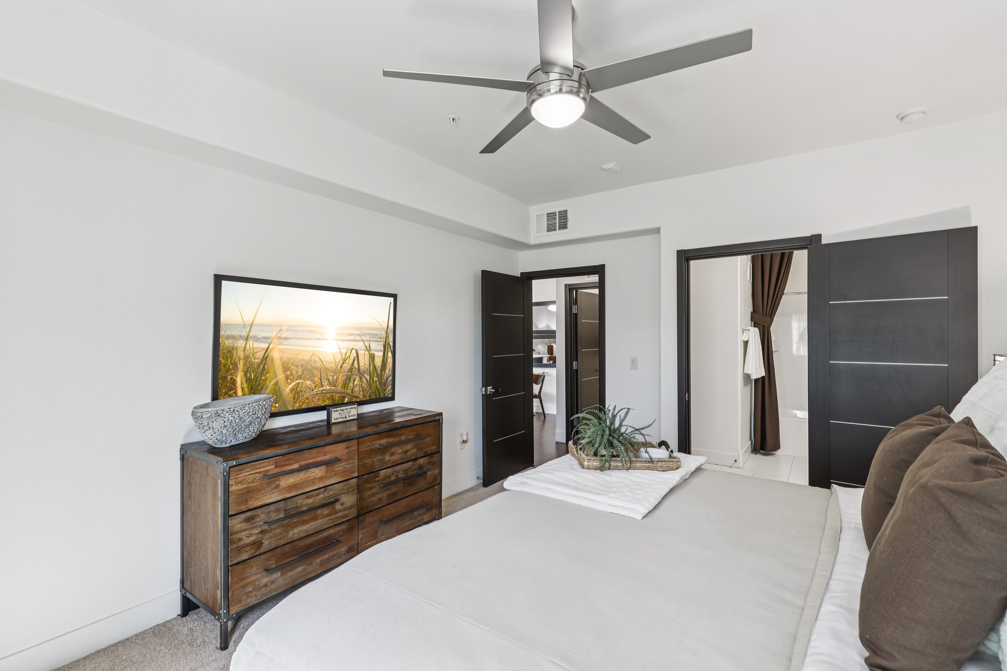 A modern bedroom featuring a king-sized bed with decorative pillows, a wooden dresser with a plant, and a flat-screen TV displaying a beach scene. The room has a ceiling fan and two partially open doors leading to a bathroom, with light-colored walls and minimalistic decor.