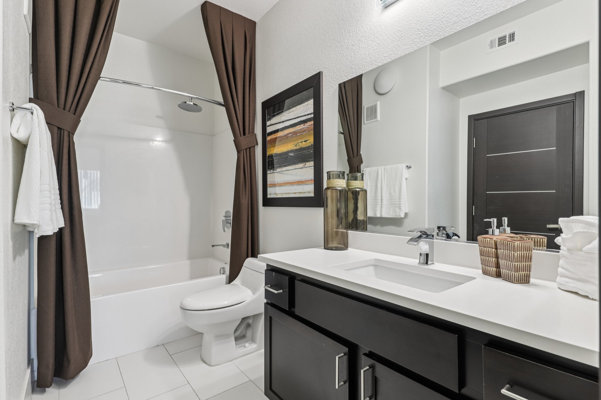 A modern bathroom featuring a bathtub with a shower, a toilet, and a stylish vanity with a sink. The decor includes dark brown curtains, a framed artwork, and a wicker basket. White towels are hanging nearby, contributing to a clean and organized look.