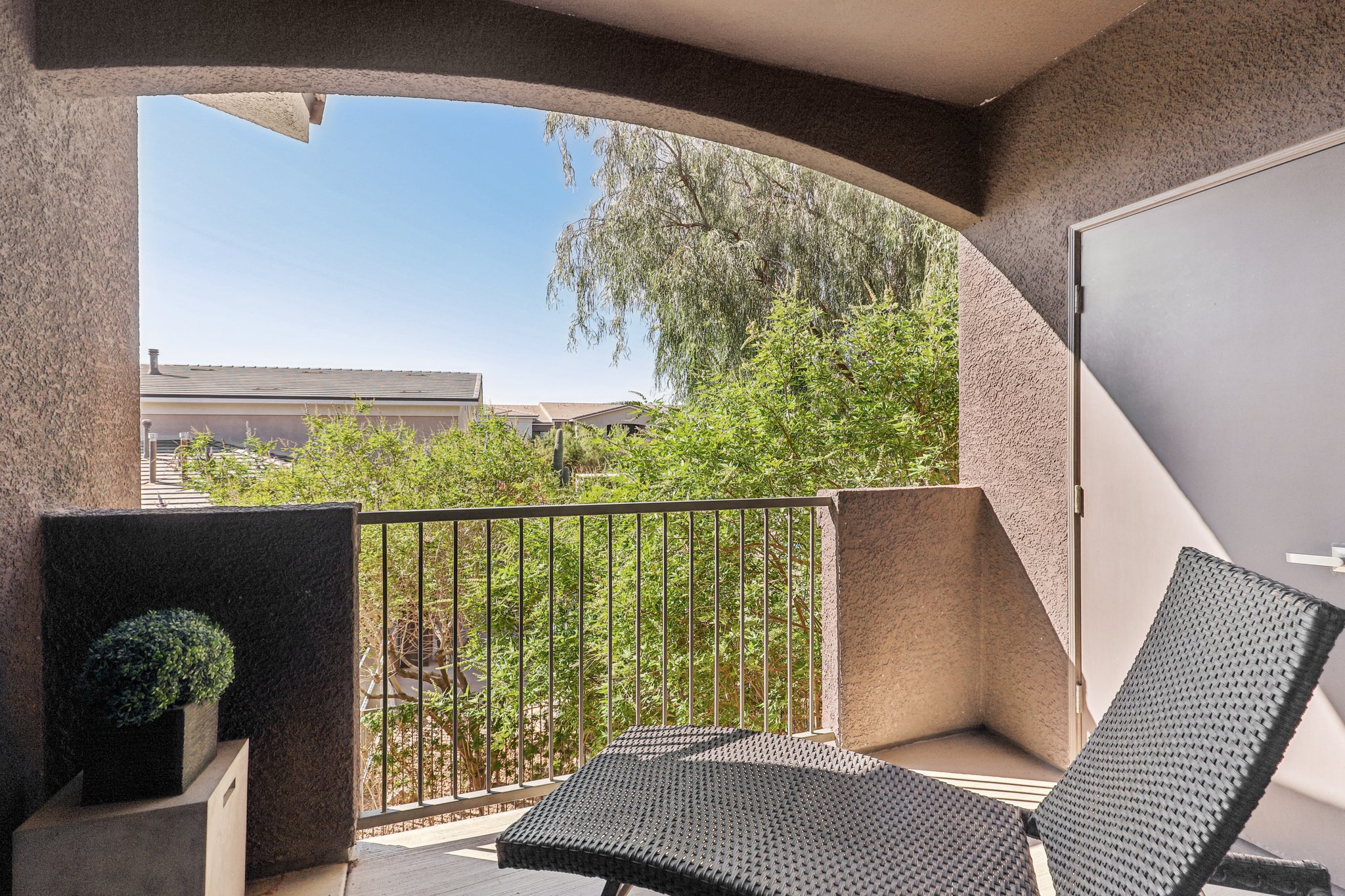 A cozy balcony with a modern black lounge chair and a small potted plant. The view features green plants and trees, creating a serene outdoor space. The railing provides a sense of enclosure while allowing light to enter. The overall atmosphere is relaxed and inviting, perfect for enjoying the outdoors.