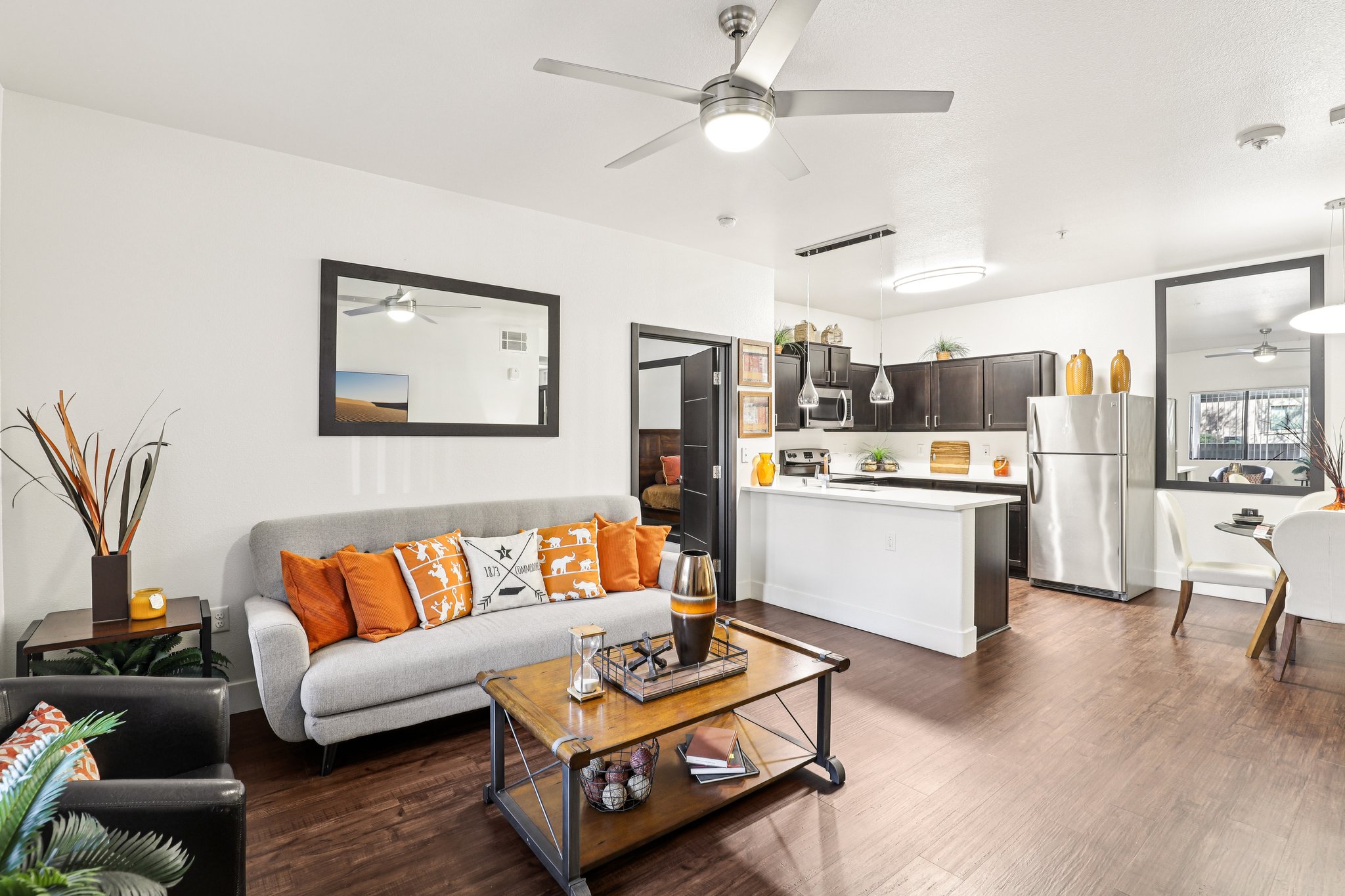 A bright and modern living room featuring a gray sofa with orange decorative pillows, a coffee table, and a view of a kitchen with dark cabinets and stainless steel appliances. The space is enhanced by stylish decor, including potted plants and wall mirrors. Wood flooring adds warmth to the inviting atmosphere.