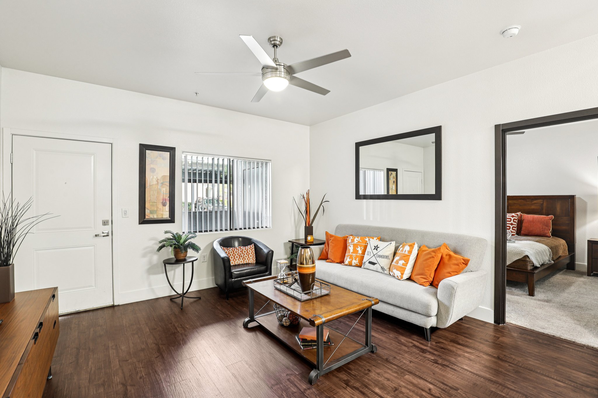 A bright, modern living room featuring a gray sofa with orange accent pillows, a wooden coffee table, and a stylish chair. The space has light wood flooring, a ceiling fan, and a decorative mirror on the wall. A door leads to another room in the background, creating a cozy and inviting atmosphere.